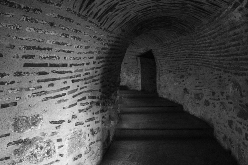 Black and white photo of a curved stone tunnel with brick walls and ascending stairs leading to a dark doorway.