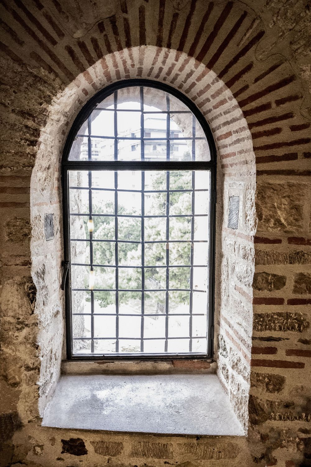 Three arched stone windows with black metal frames in an ancient monastery wall show views of green trees outside.