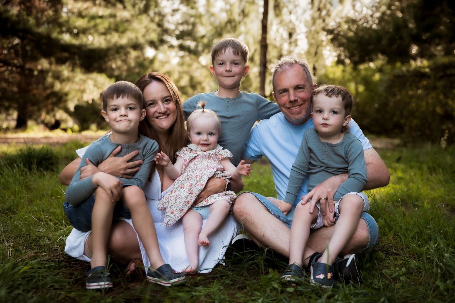 A family of six wearing coordinating blue outfits sits together on grass during a sunny outdoor photo session.