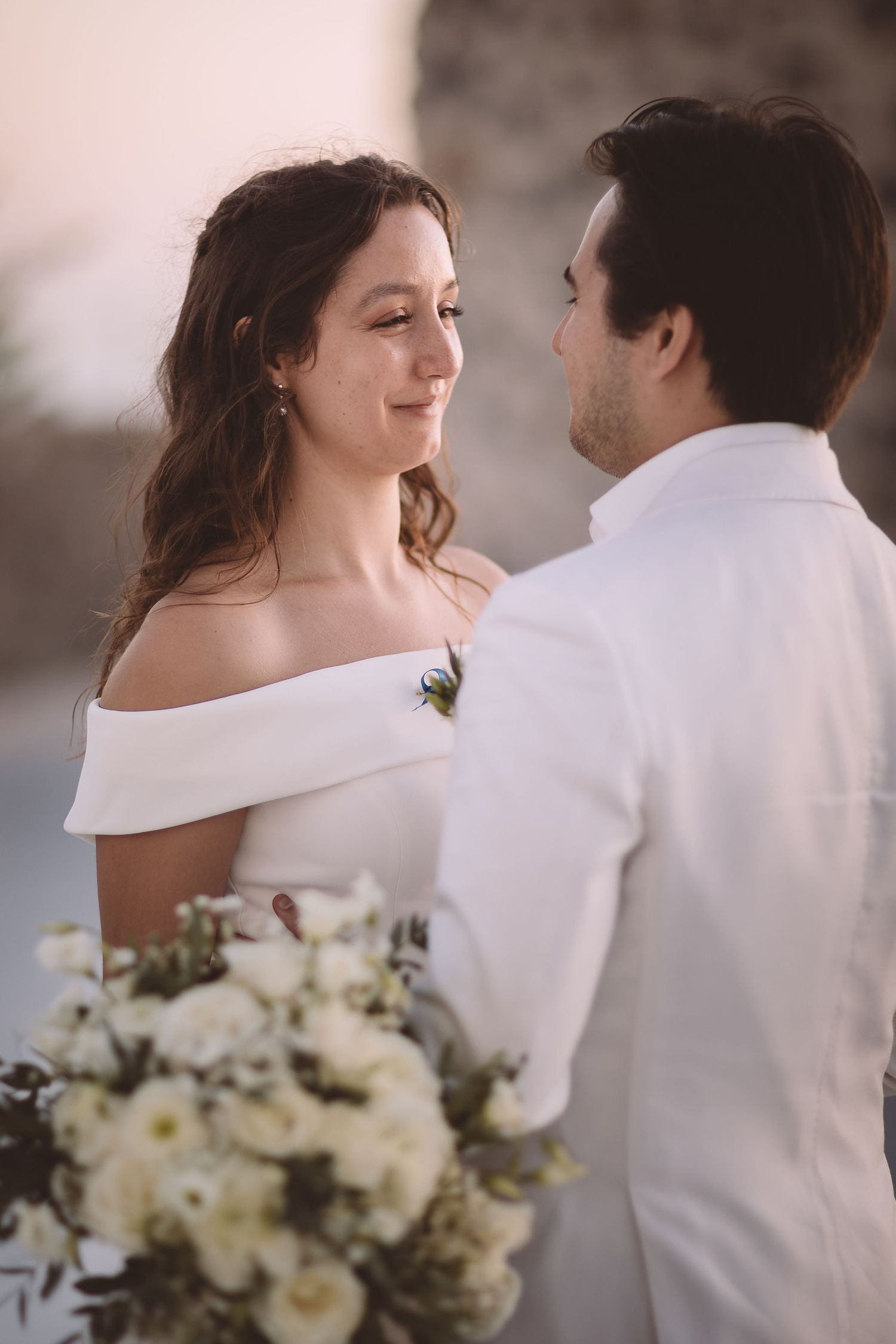 Destination wedding couple embraces in soft light at El Viento, Santorini with elegant white bouquet of flowers and matching attire.