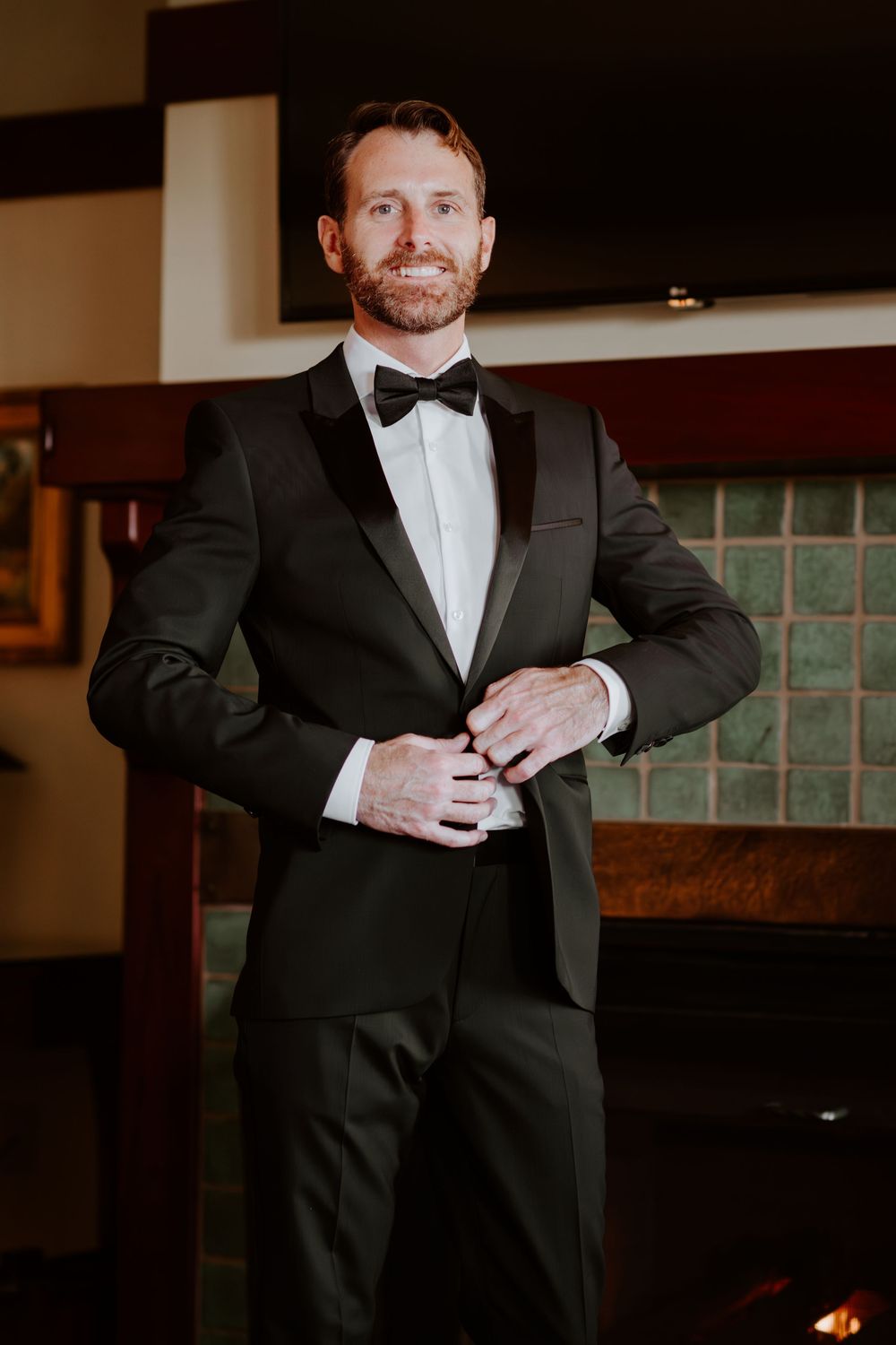 Handsome groom in black tuxedo buttoning jacket while smiling near a fireplace.