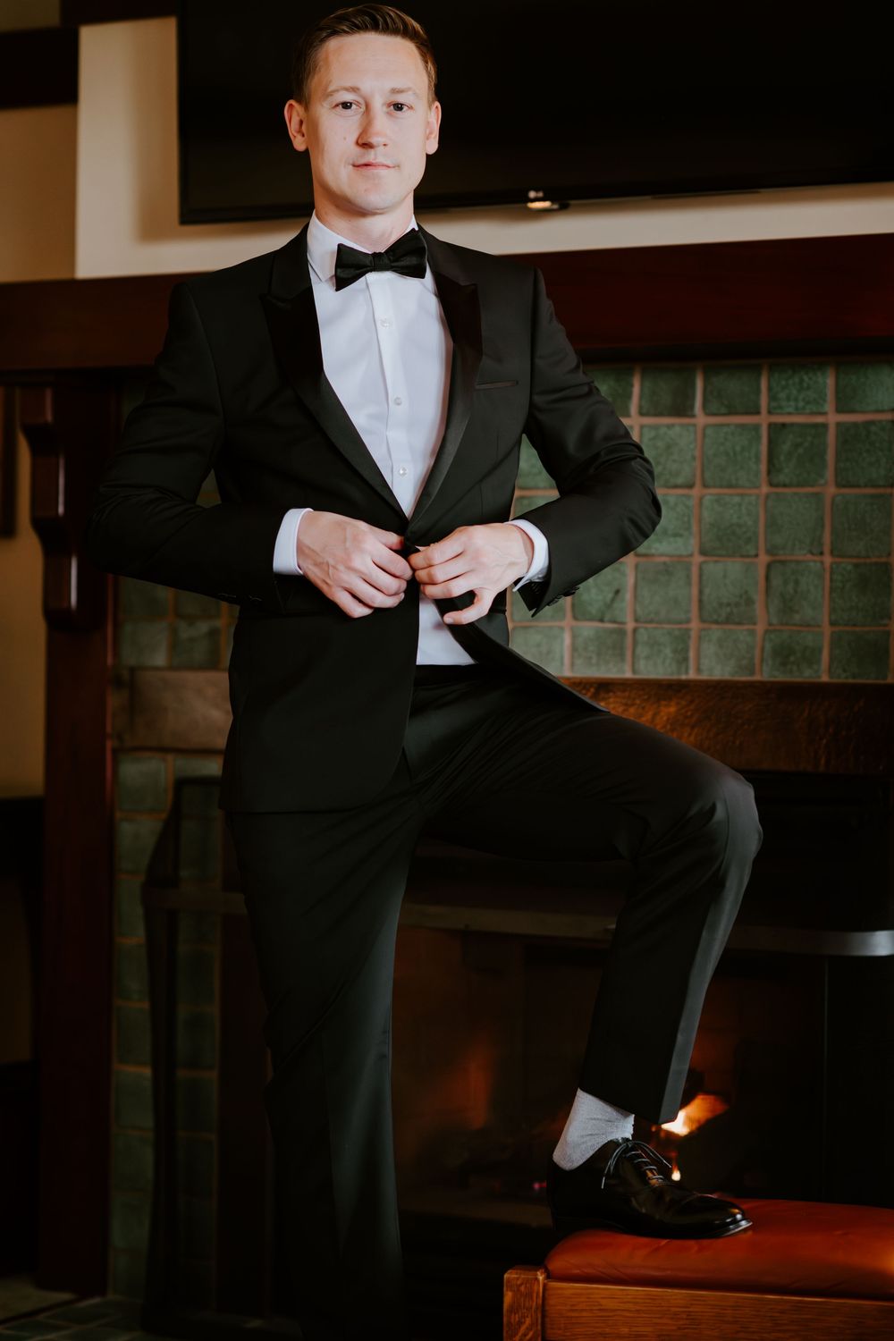 Groom in black tuxedo buttoning jacket with one foot raised on a bench near fireplace.