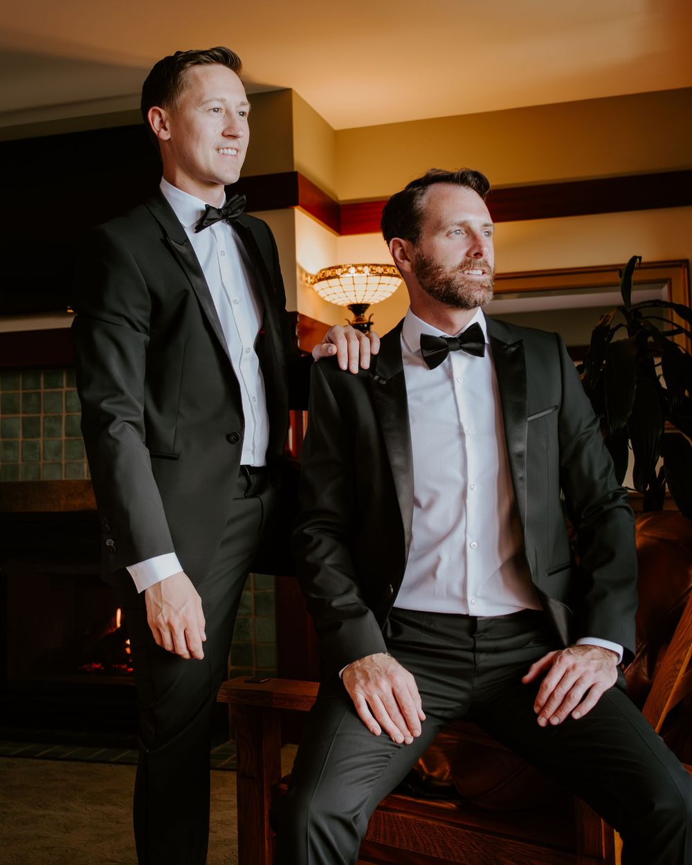 Two groomsmen in black tuxedos posing together near a warmly lit fireplace indoors.