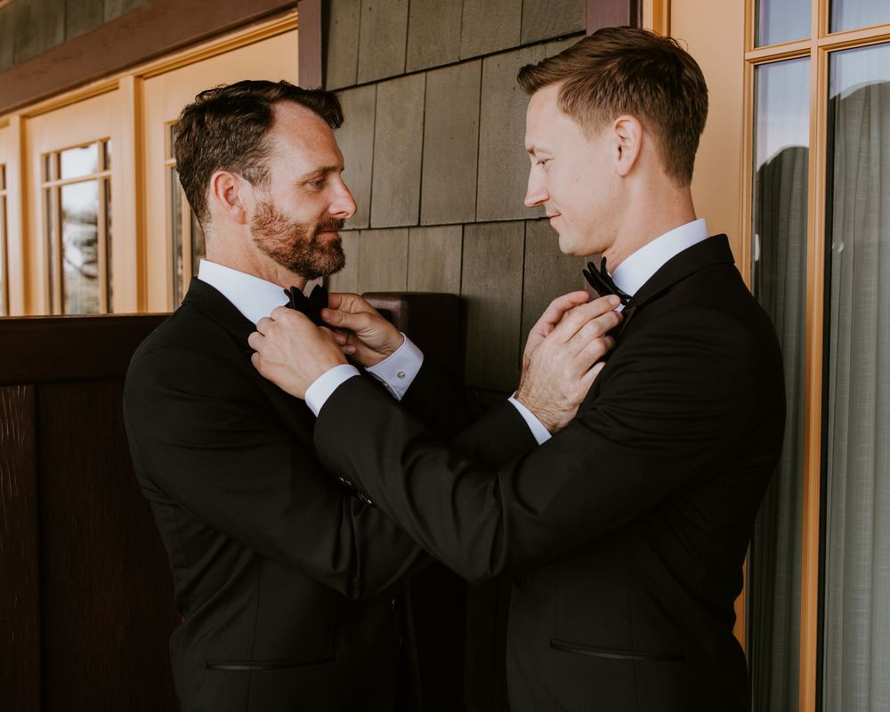 Two grooms in black tuxedos adjusting each other's bow ties outdoors before wedding ceremony.