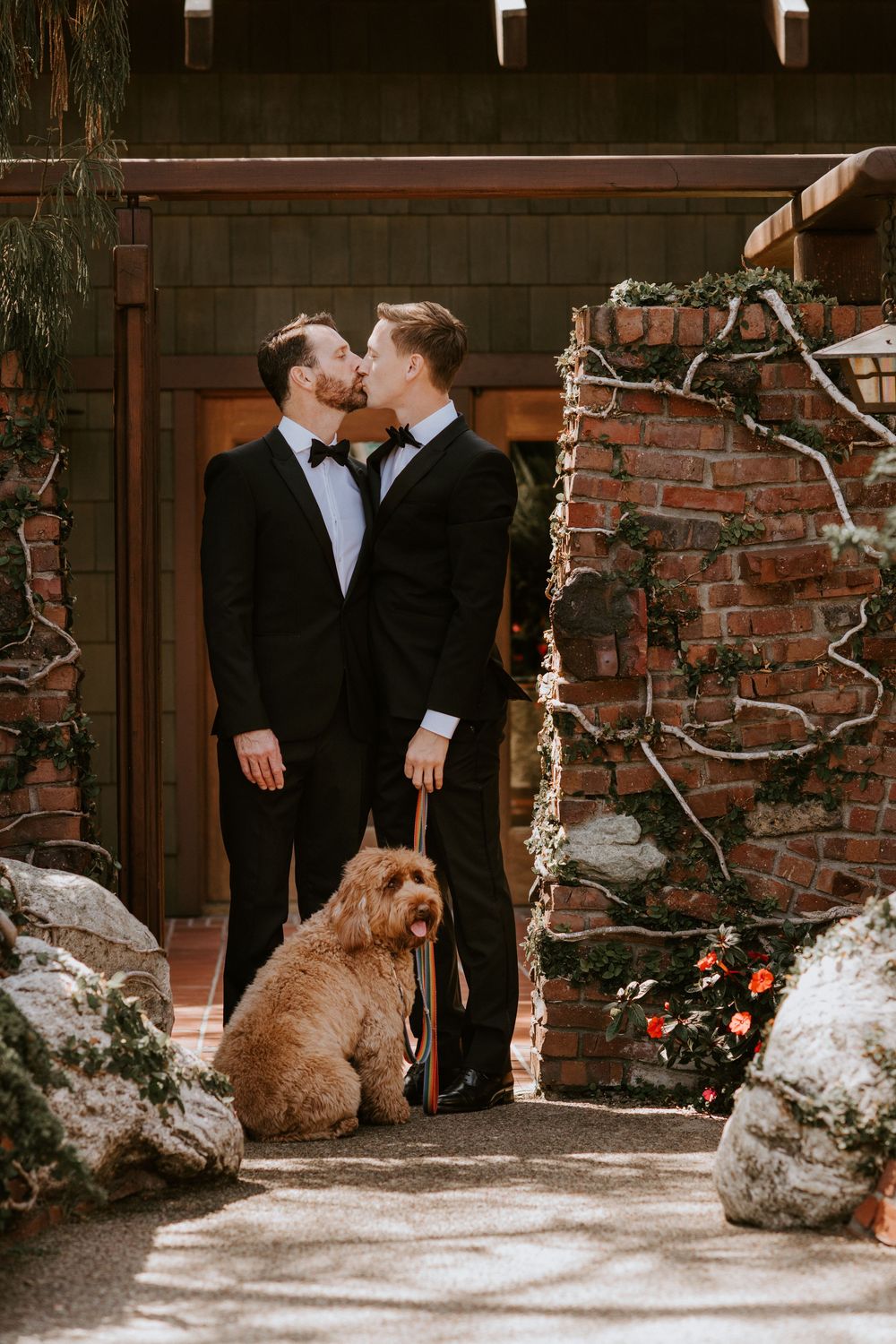 Two grooms in black tuxedos share a kiss outside a rustic brick venue with their golden doodle dog.