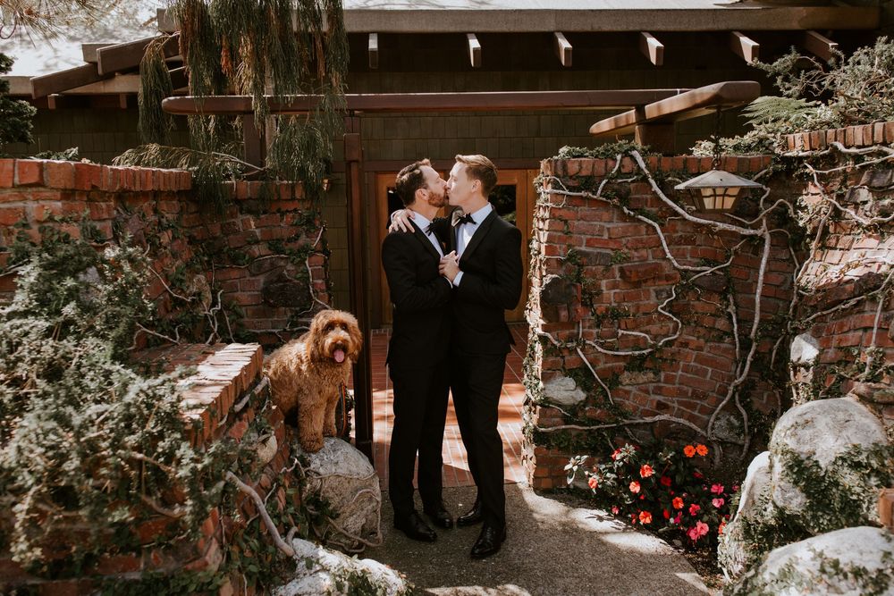 Same-sex couple in matching black tuxedos embrace romantically outside a rustic brick and stone wedding venue.