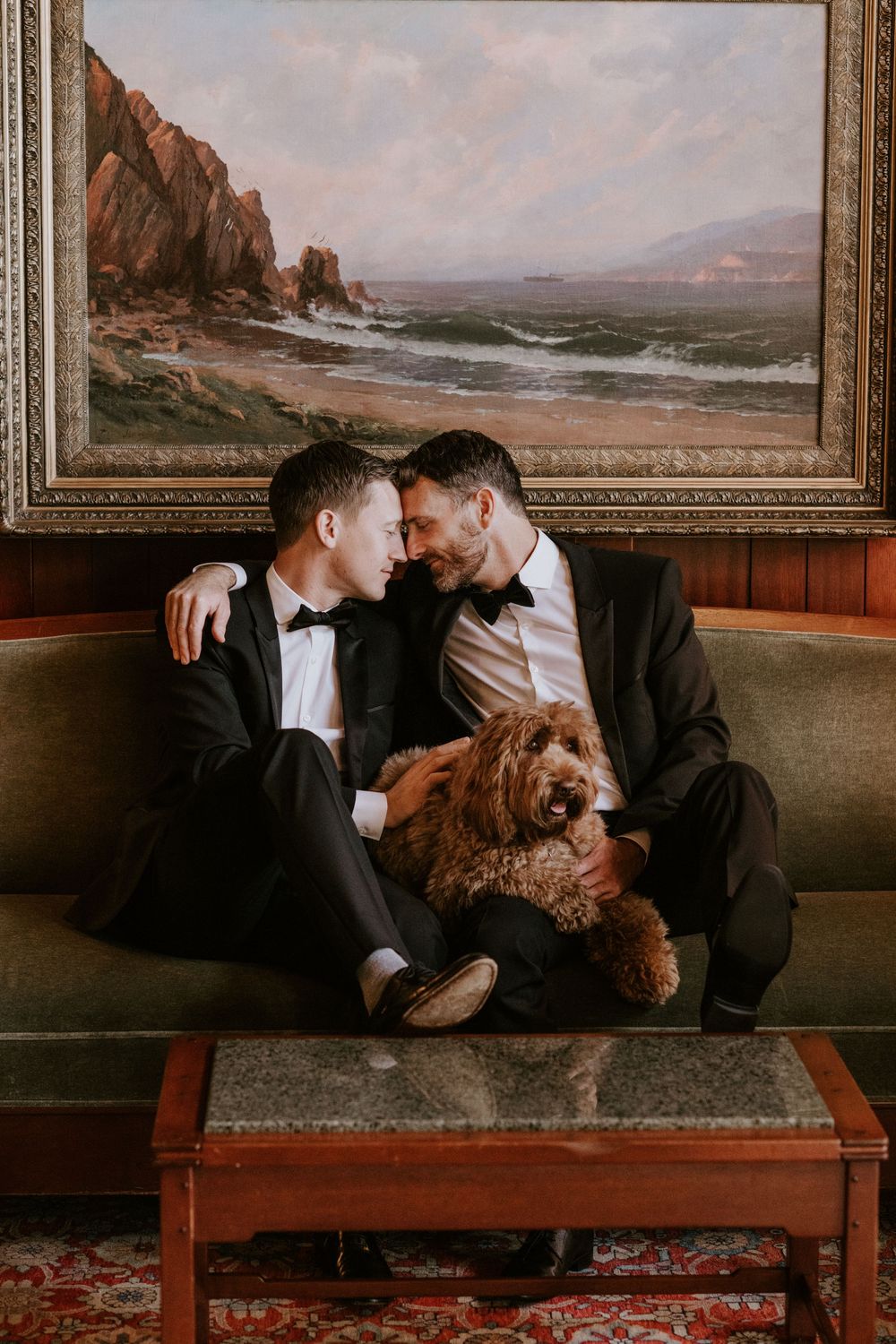 Two grooms in black tuxedos sit on a vintage sofa with their golden doodle dog beneath a coastal painting.