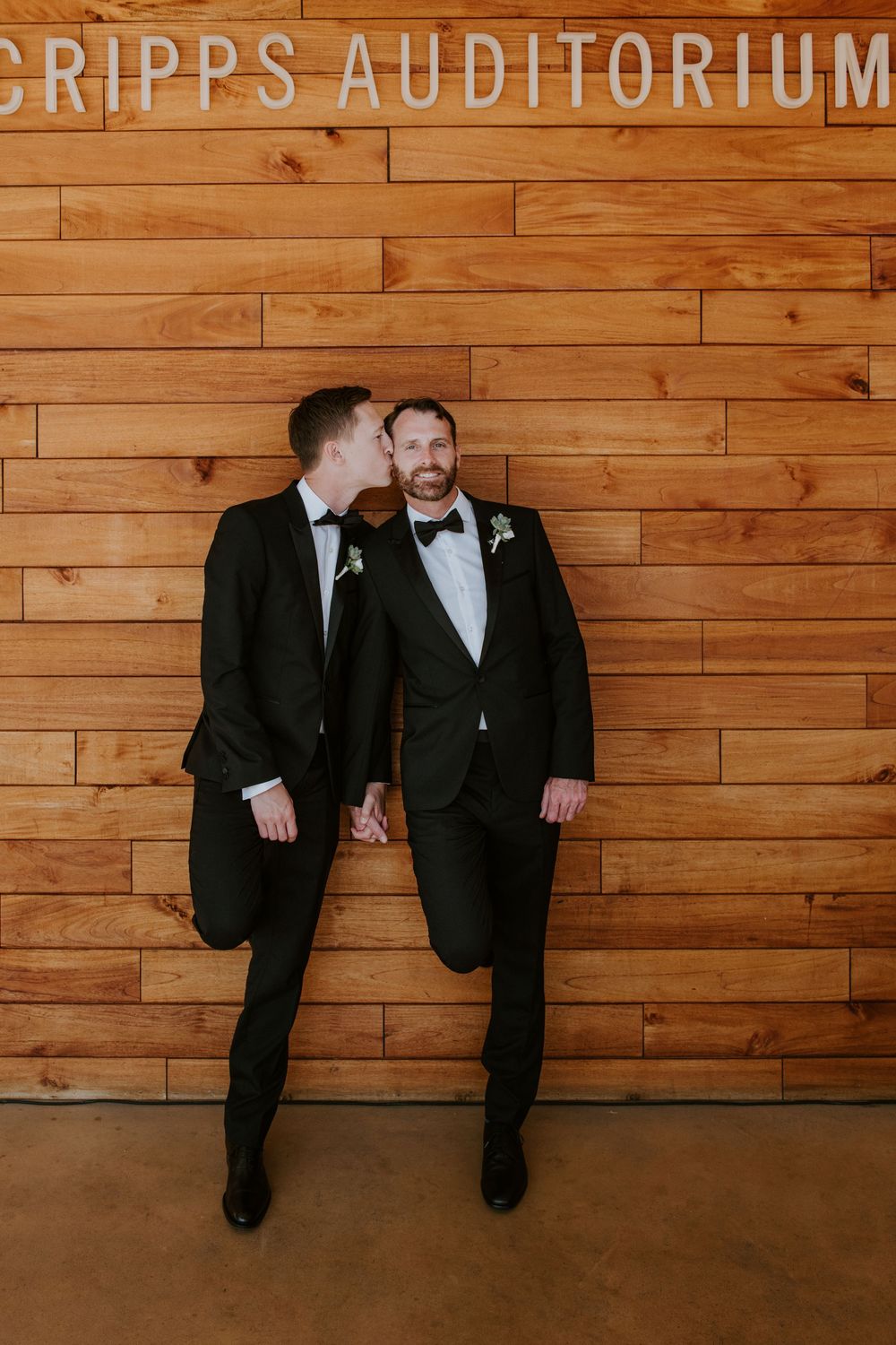 Two grooms in black tuxedos hold hands and share a kiss outside Scripps Auditorium wooden wall backdrop.