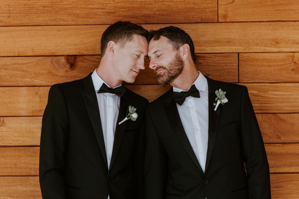 Two grooms in black tuxedos with boutonnieres touch foreheads tenderly against a warm wooden wall.