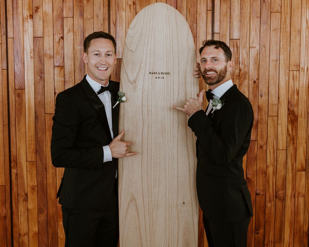 Two grooms in black tuxedos smile proudly beside a personalized wooden surfboard at their wedding reception.