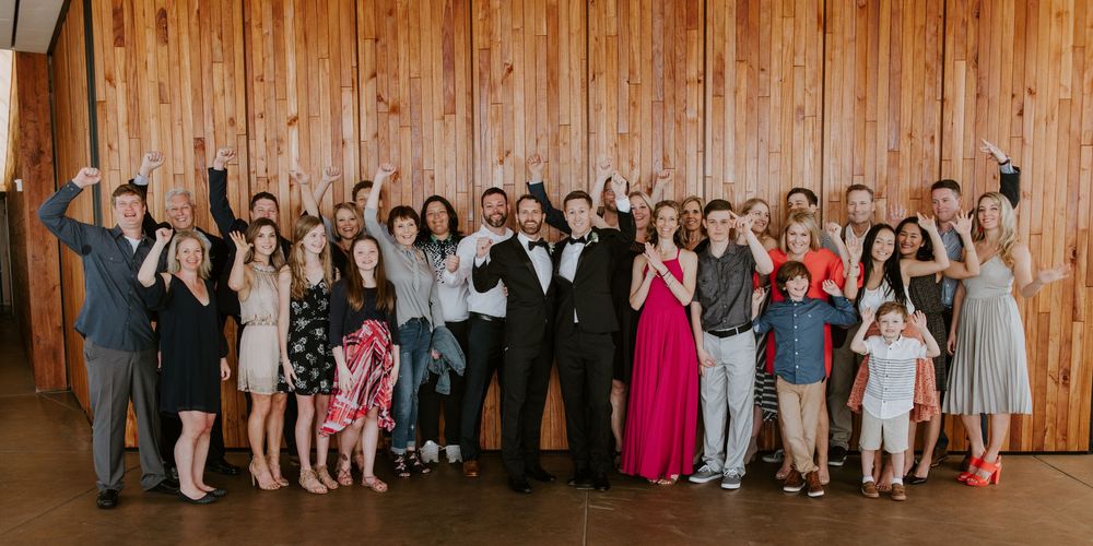 Large wedding party group celebrates joyfully with raised hands in a warm wood-paneled reception venue.