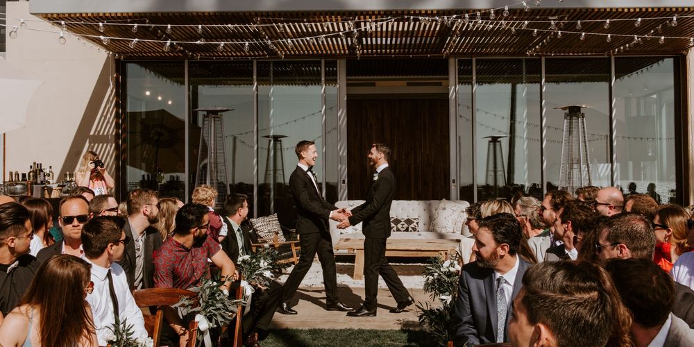 Two grooms in black tuxedos exchange vows at an outdoor wedding ceremony surrounded by seated guests.