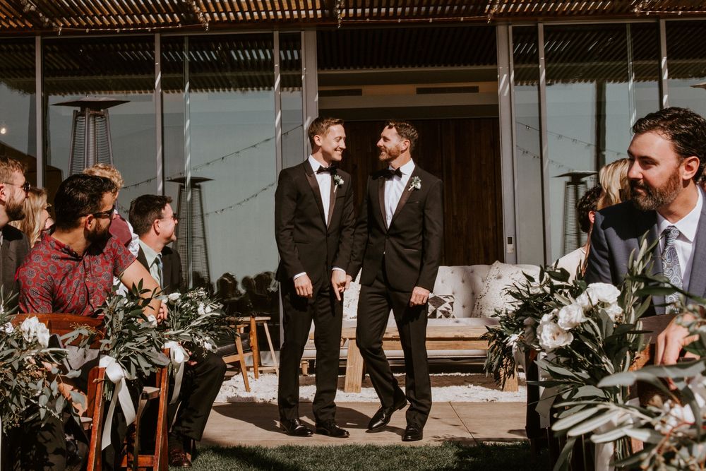 Two grooms in black tuxedos smile and walk the aisle at their outdoor wedding ceremony with guests.