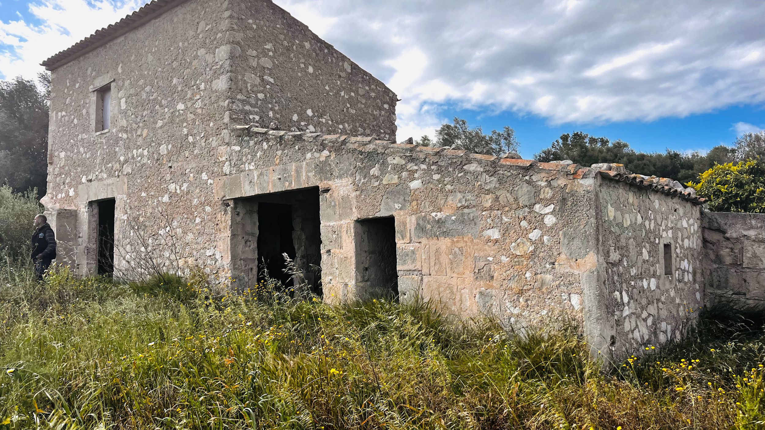 An old, stone building with a dilapidated exterior stands in a grassy field against a cloudy sky.