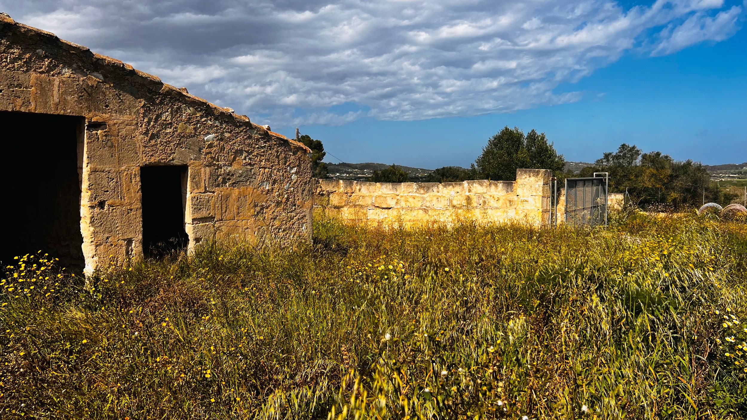 A rustic stone building sits in a grassy field against a cloudy sky backdrop.
