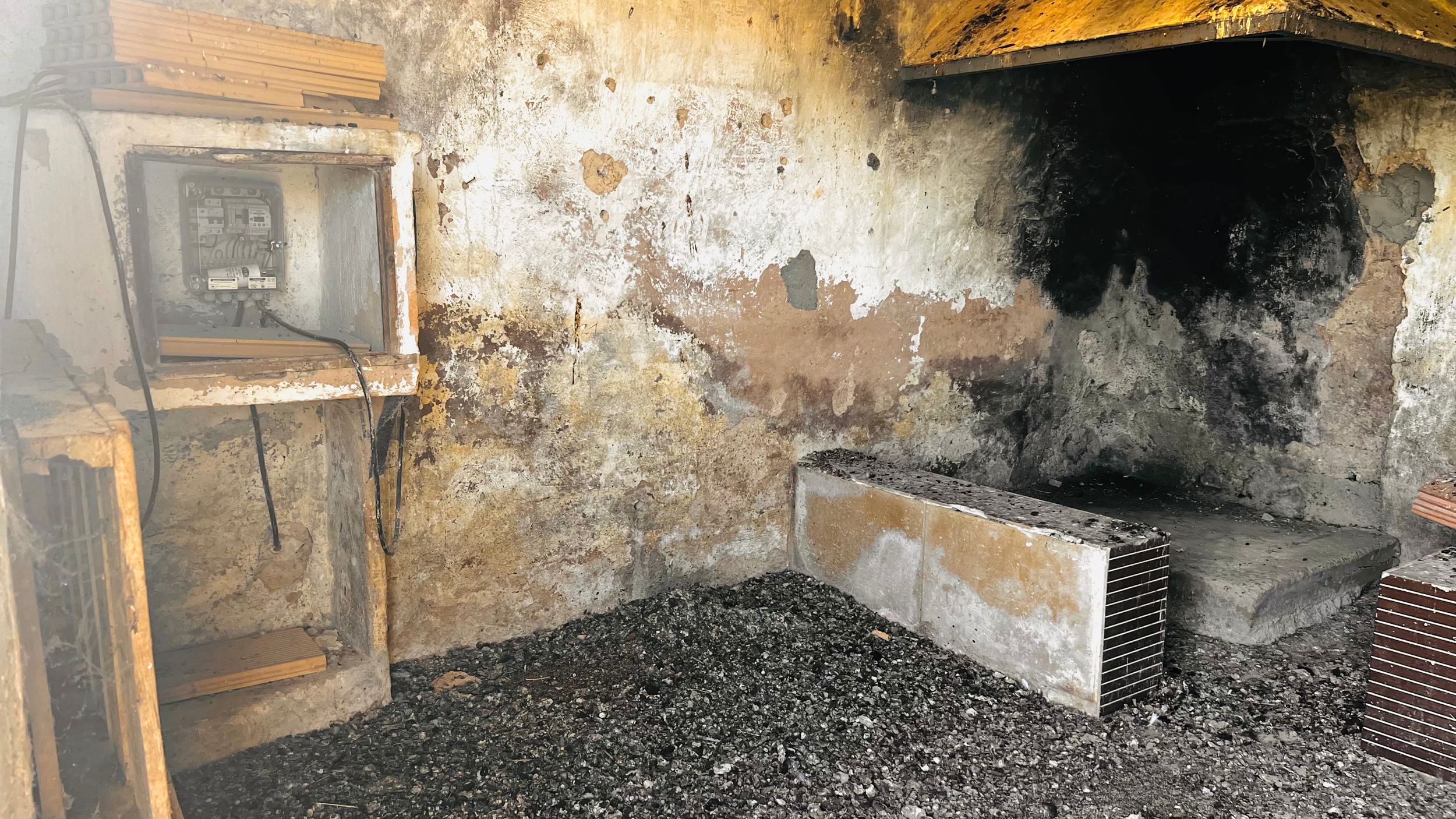 An old, rustic fireplace with a large stone hearth and piles of firewood in a dimly lit room.