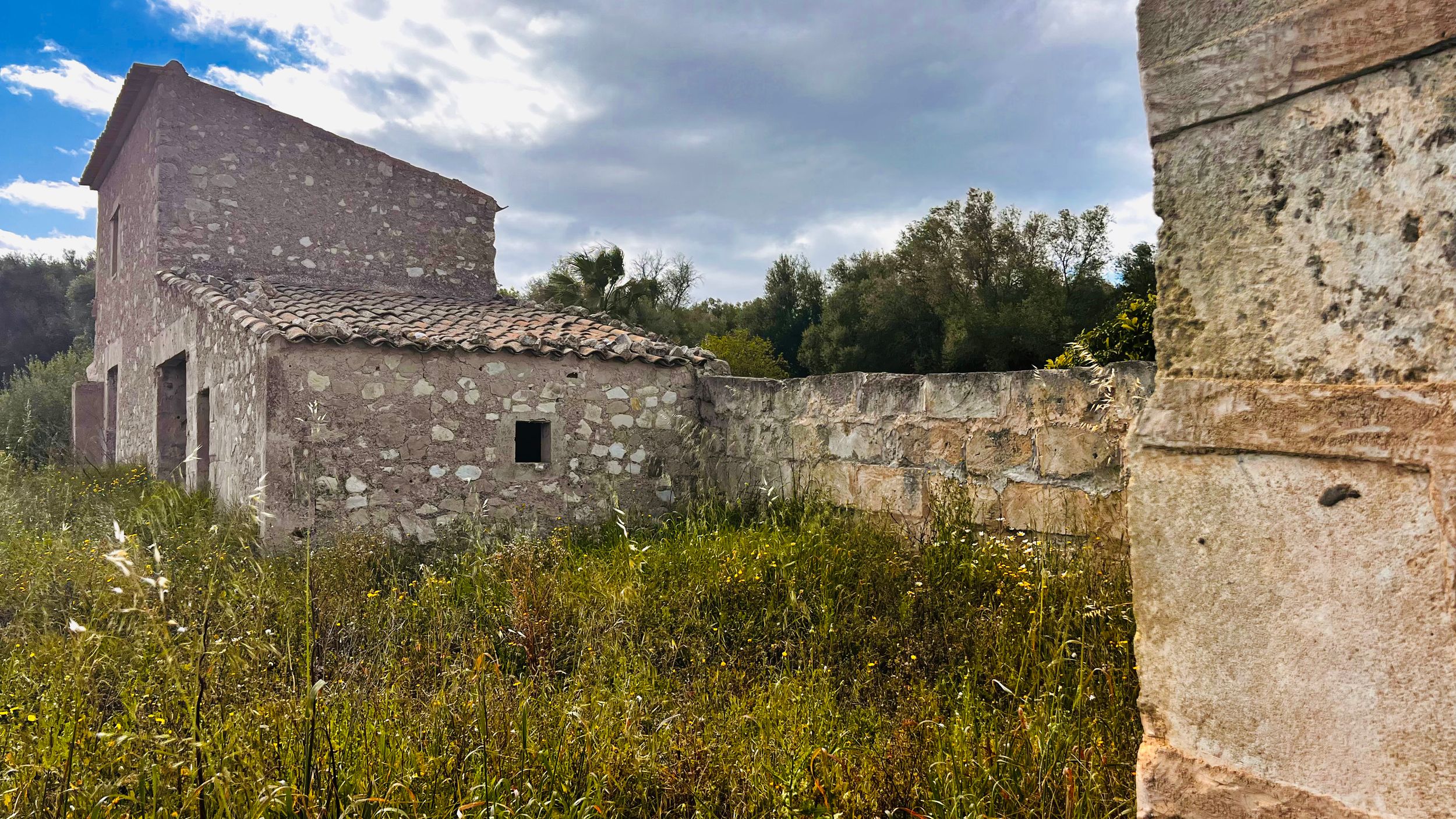 An old stone building surrounded by overgrown vegetation and a cloudy sky in the background.
