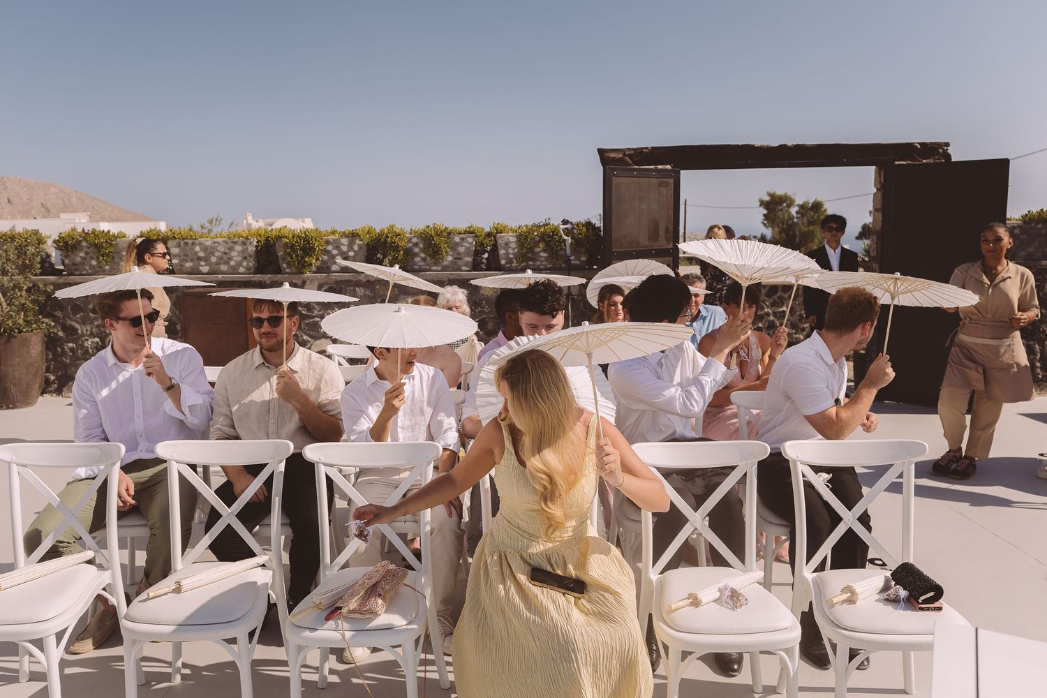 A group of wedding guests at El Viento Santorini are seated at an outdoor wedding holding parasols.