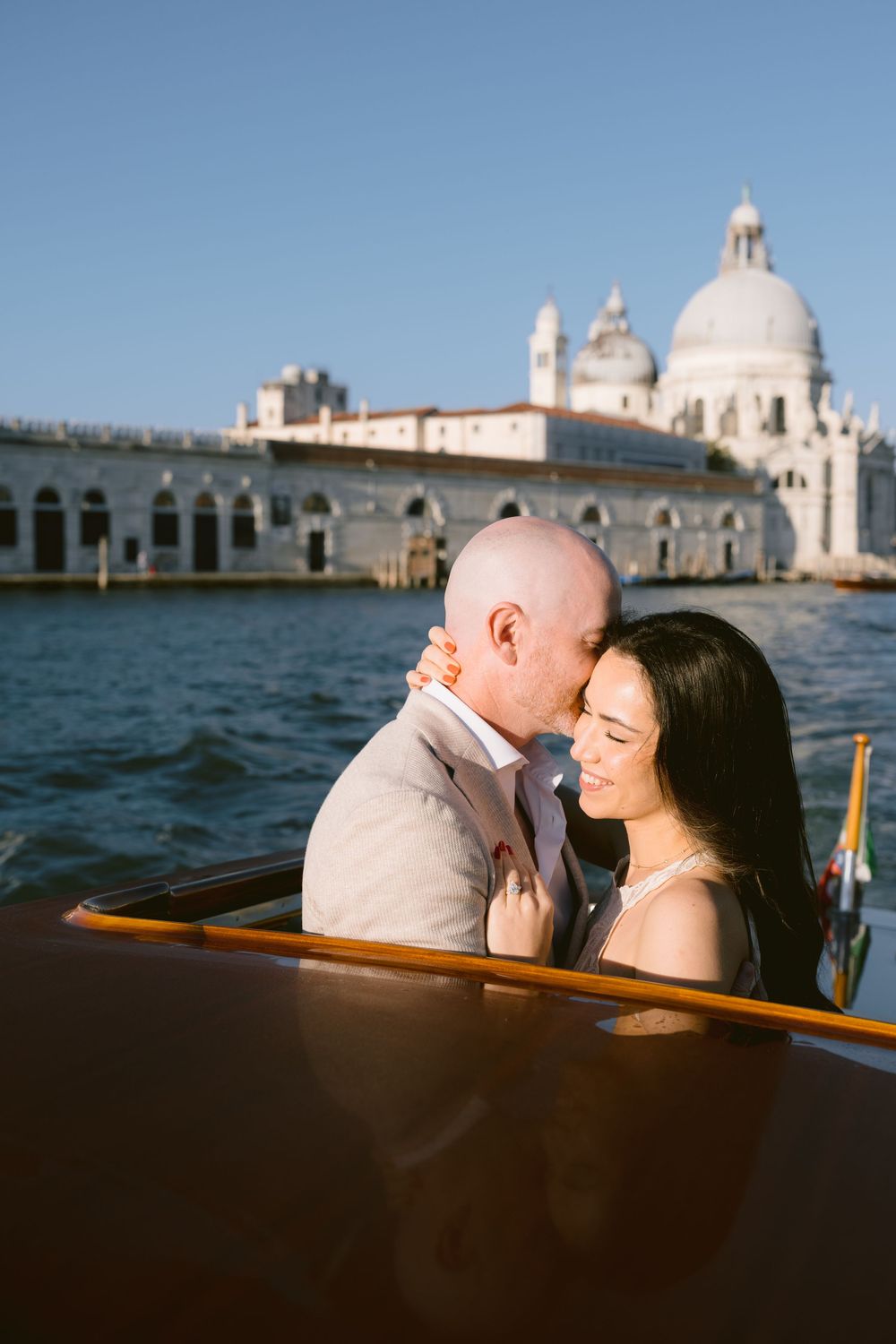 Couple sharing romantic kisses on a boat ride in Venice with the iconic domed architecture visible across the water.
