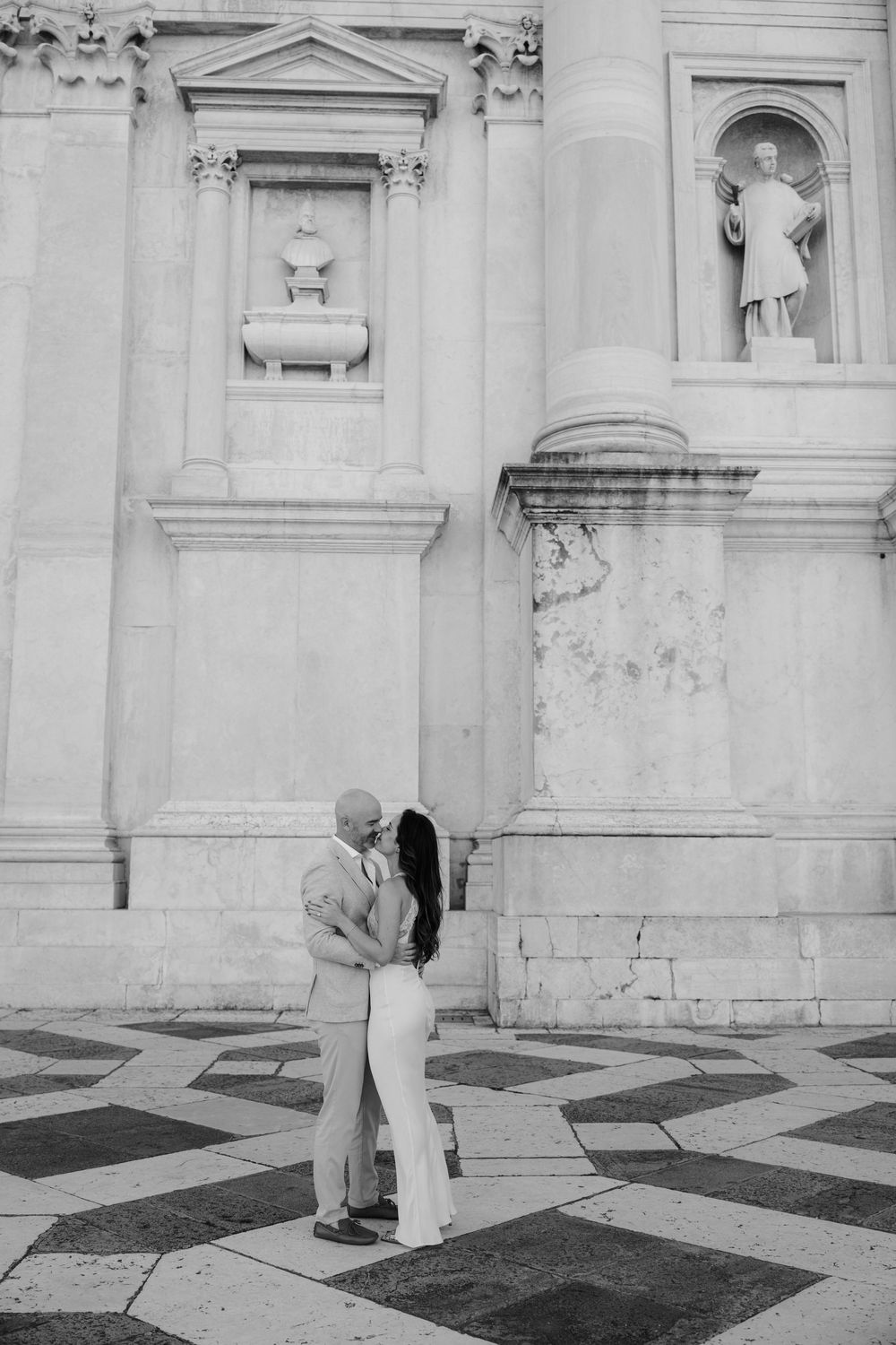 A romantic black and white sequence of a couple dancing on a patterned stone floor in front of a classical building.