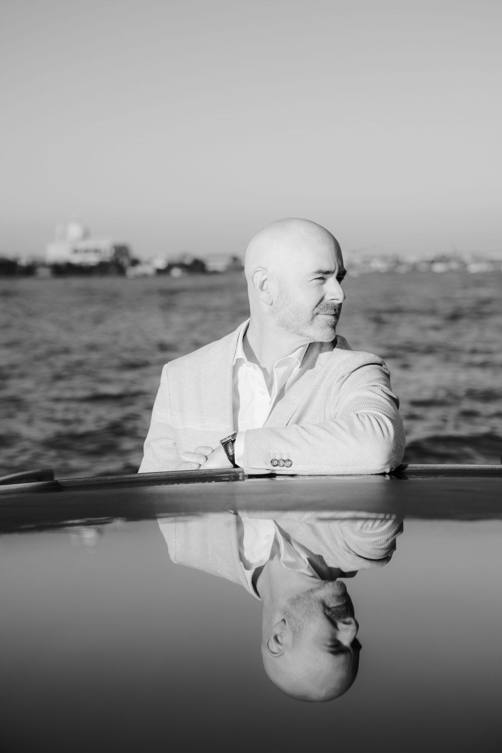 Black and white portrait of person in white suit jacket reflected in water surface with boats in background.