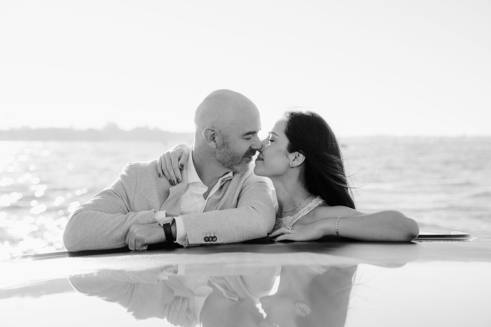 A romantic black and white photo of a couple sharing a tender kiss while leaning on a surface near the ocean.