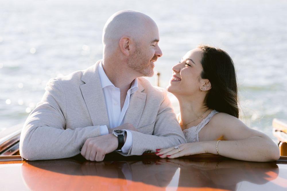 A couple shares a joyful moment while leaning on a wooden surface near the ocean during sunset.