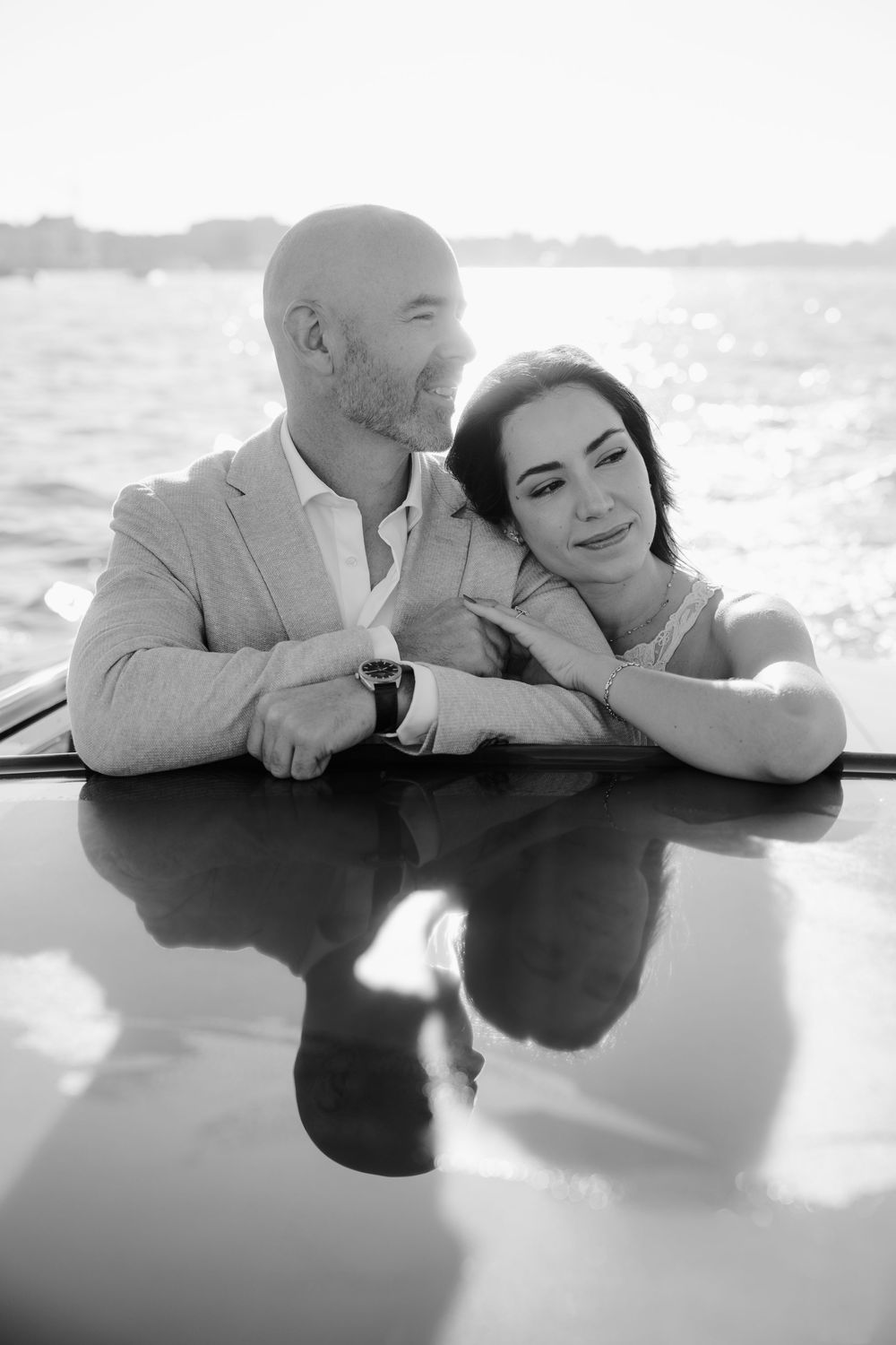A romantic black and white photo of a couple sharing an intimate moment while leaning on a reflective surface near water.