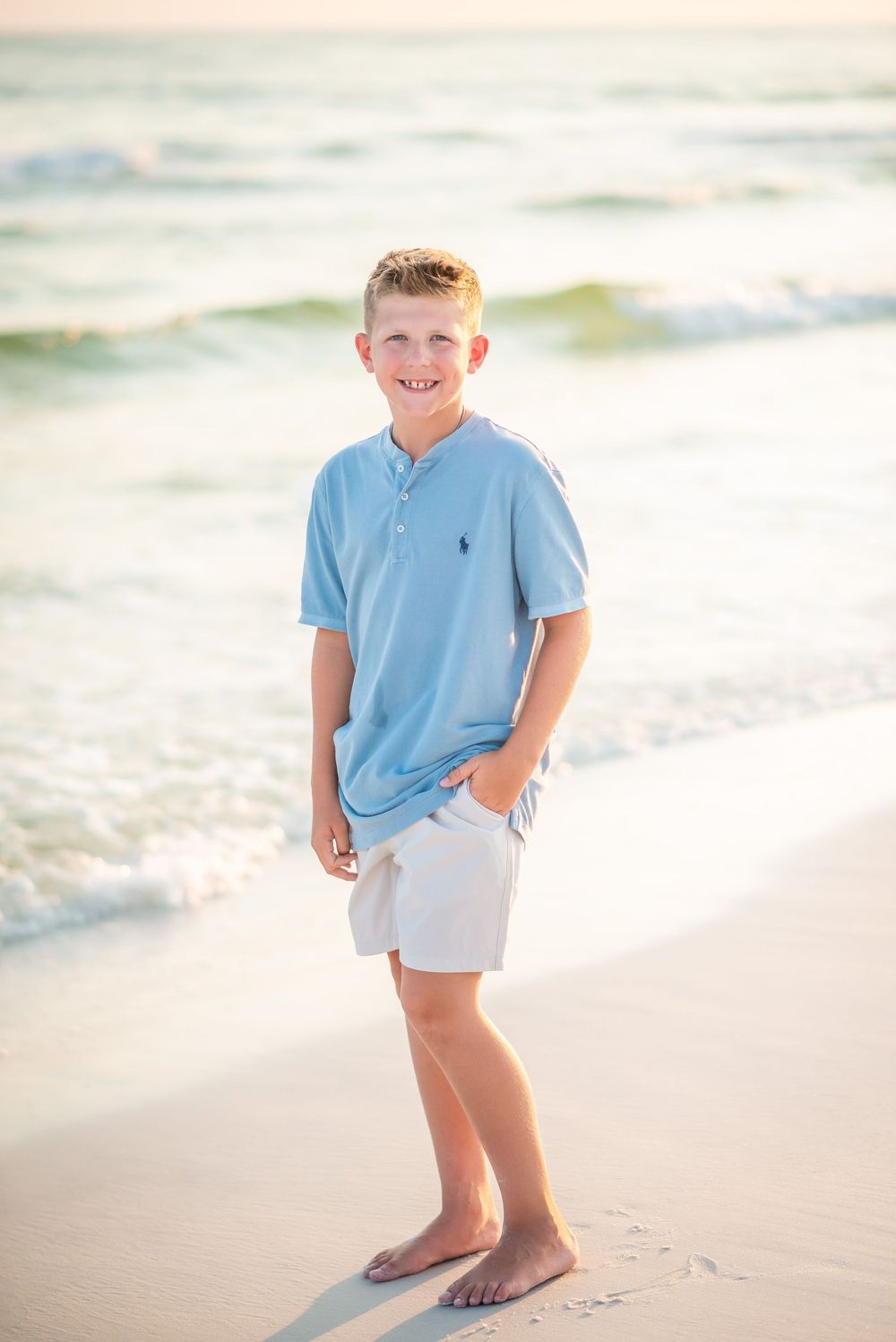 Young person in light blue polo and white shorts walking on a sunny beach at sunset.