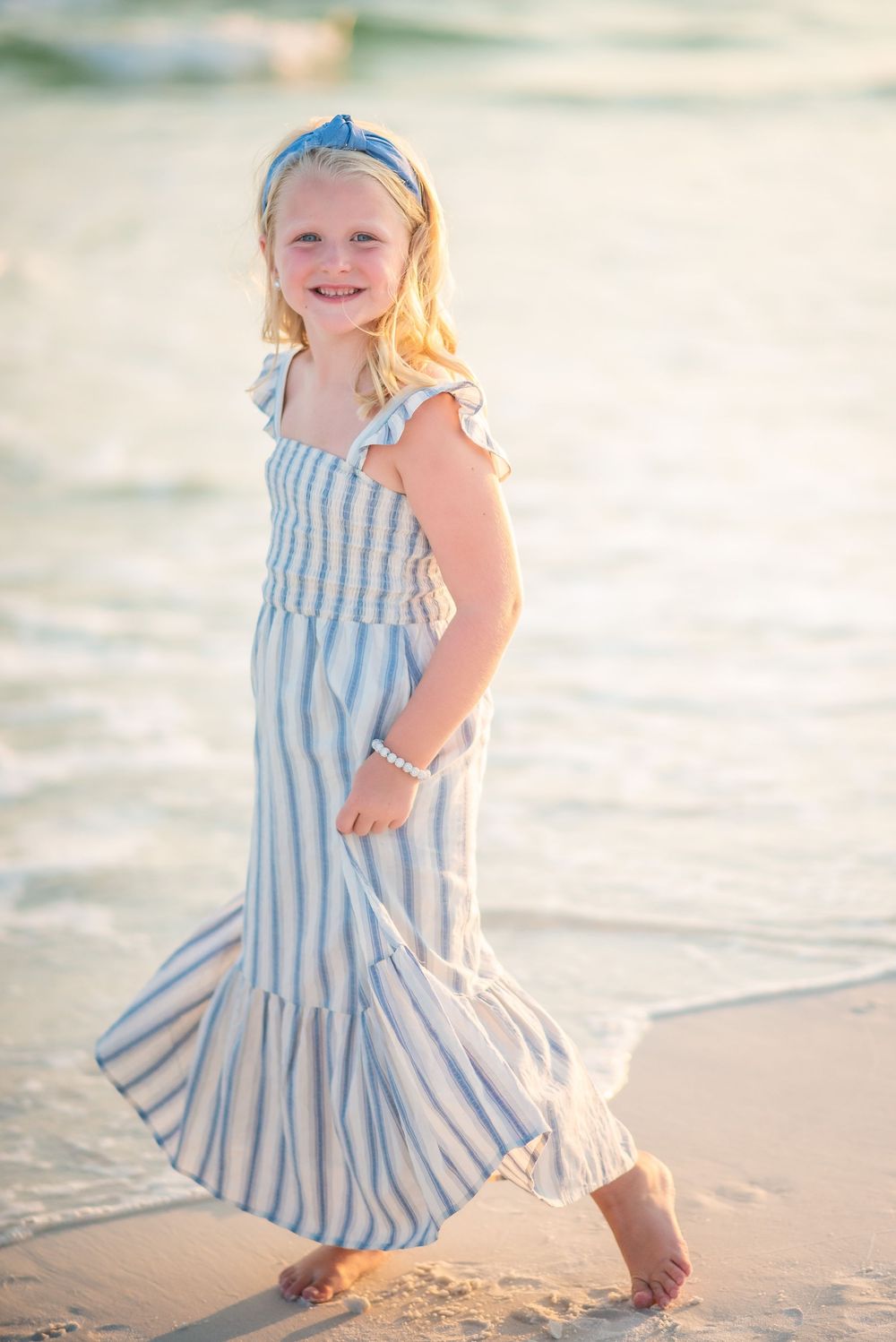 A young person in a blue and white striped dress twirls on the beach at sunset.