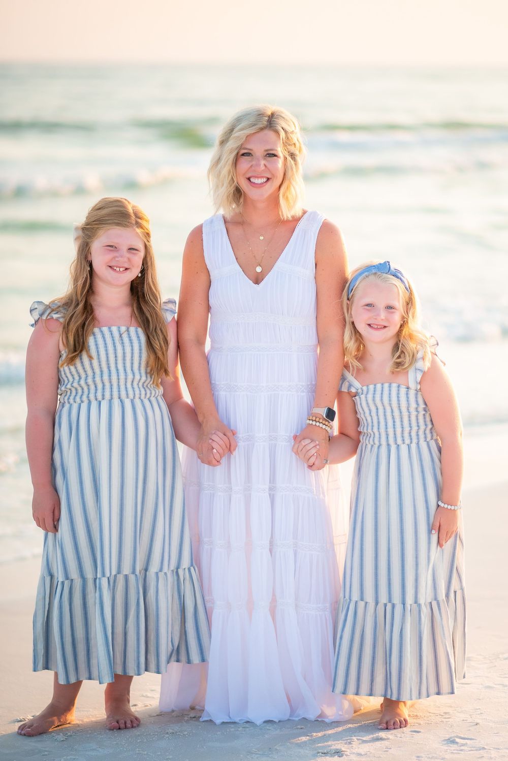 Family wearing coordinated white and blue outfits pose together on a sunny beach at sunset.