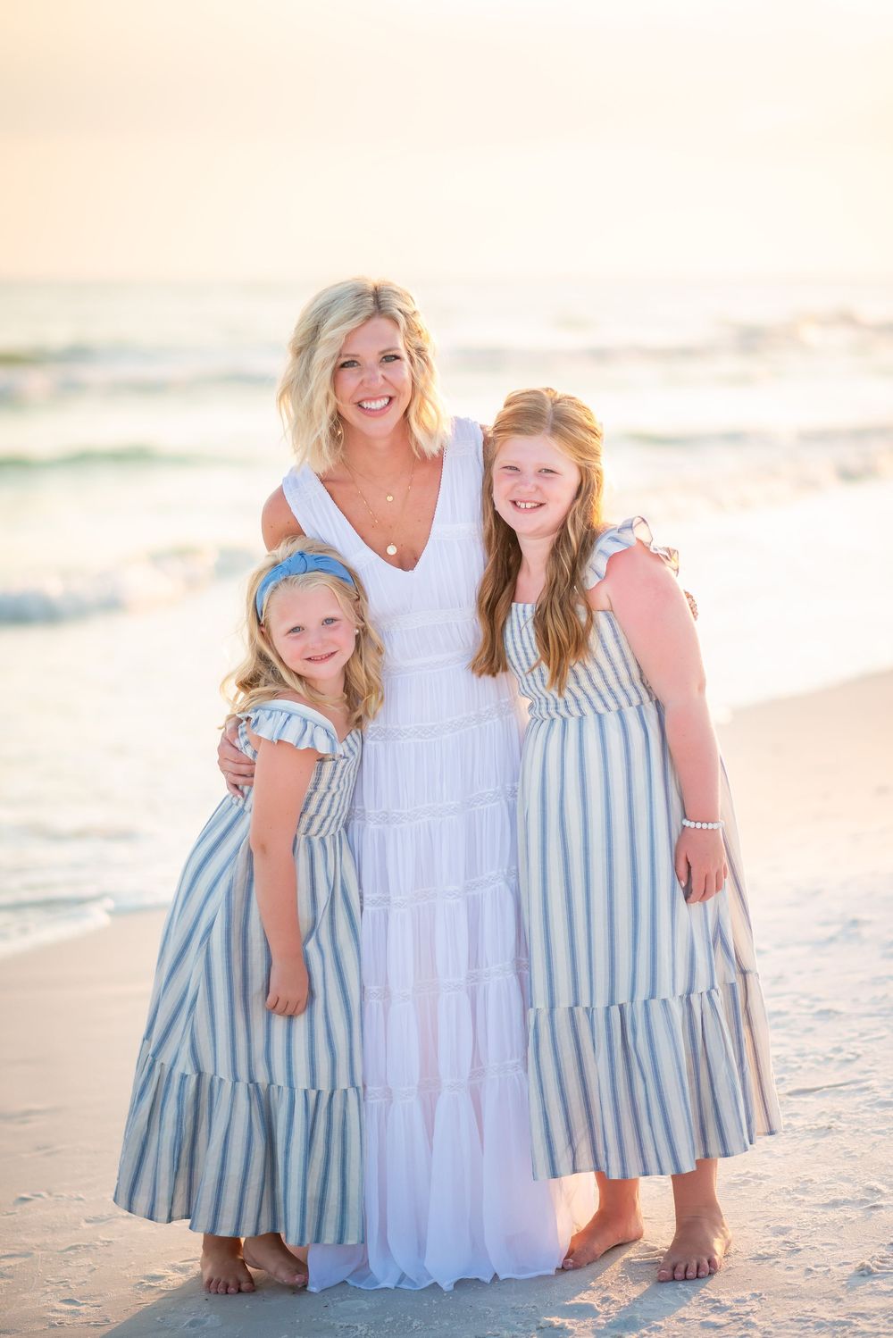 Three people in coordinating dresses pose together on a beach at sunset, wearing white and blue striped outfits.