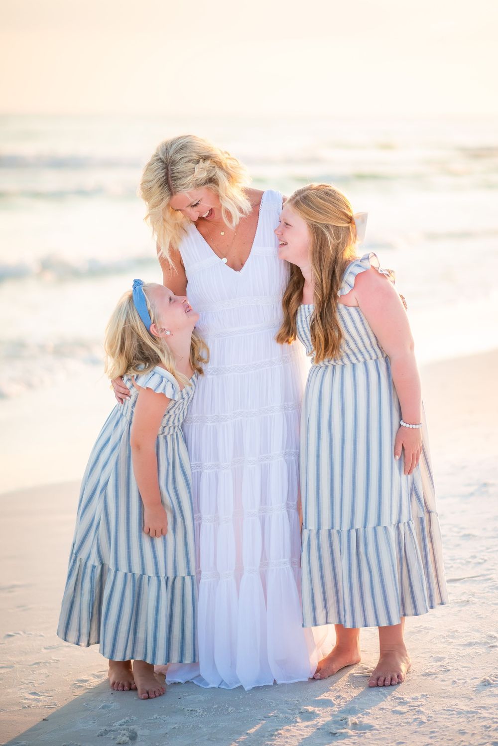 Three people in coordinating white and blue striped dresses share a joyful moment together on a sunset beach.