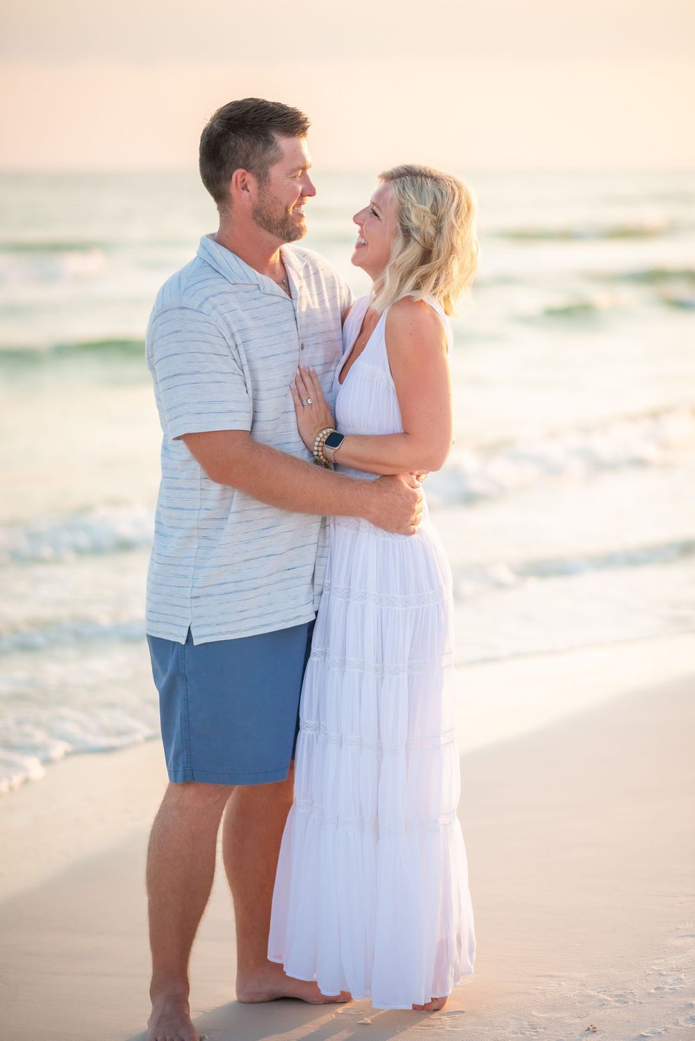 Romantic couple embracing on sandy beach at sunset wearing white and blue casual attire.