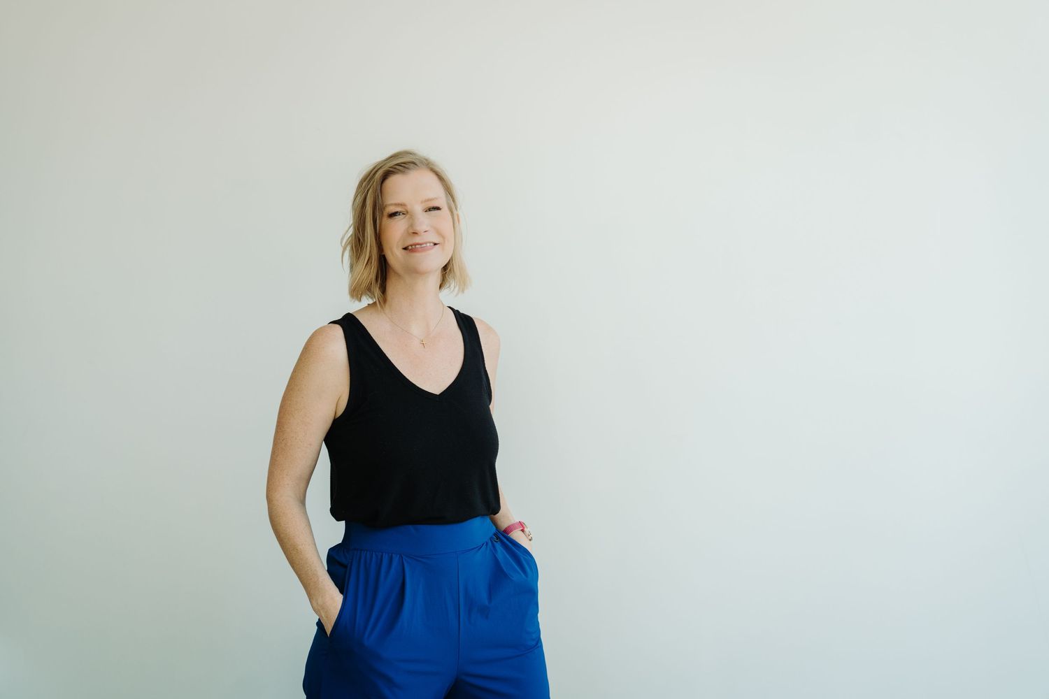Someone wearing a black sleeveless top and bright blue skirt stands against a white wall with hands in pockets.