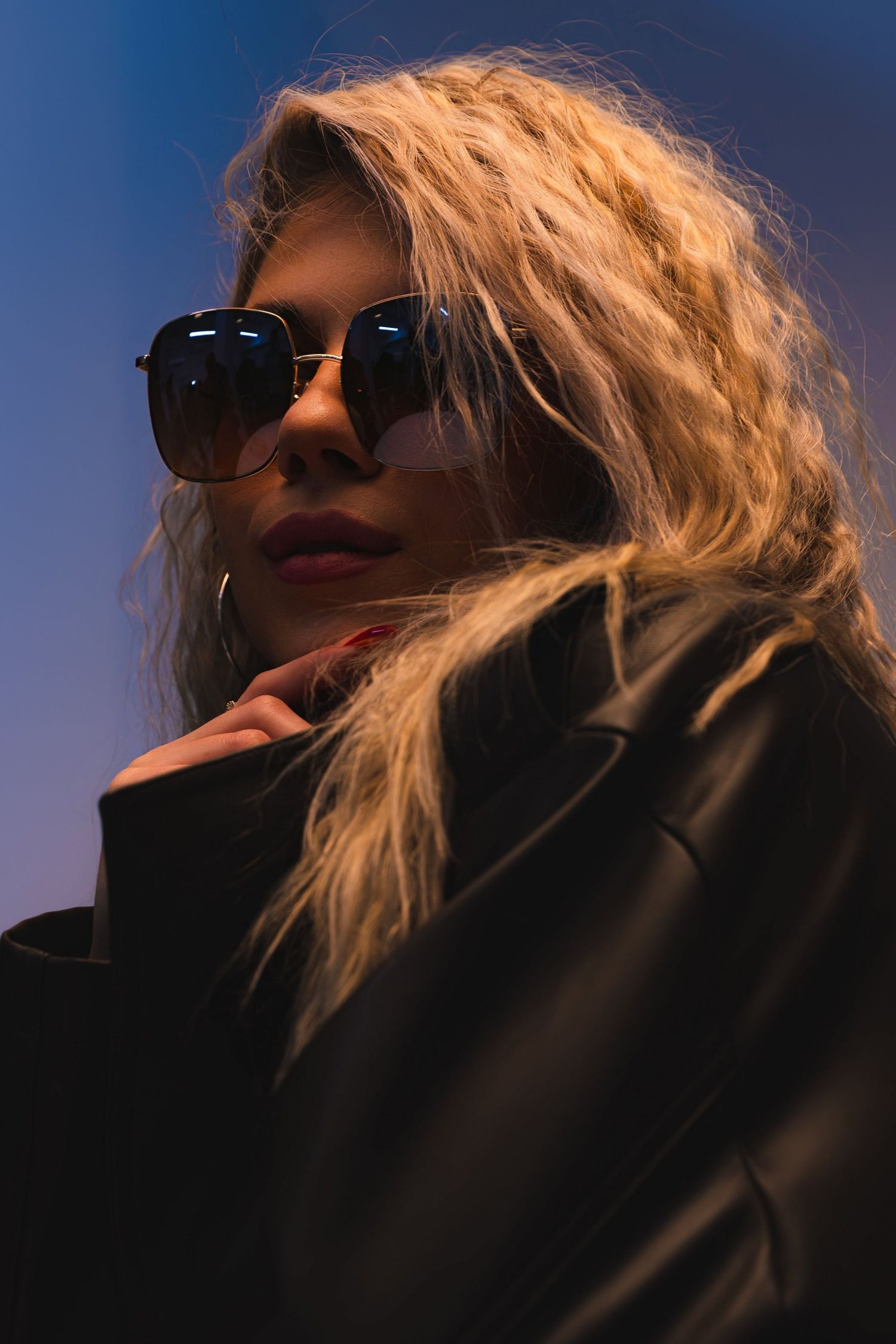 Blonde woman in sunglasses and black leather jacket posed under dramatic blue and purple lighting.