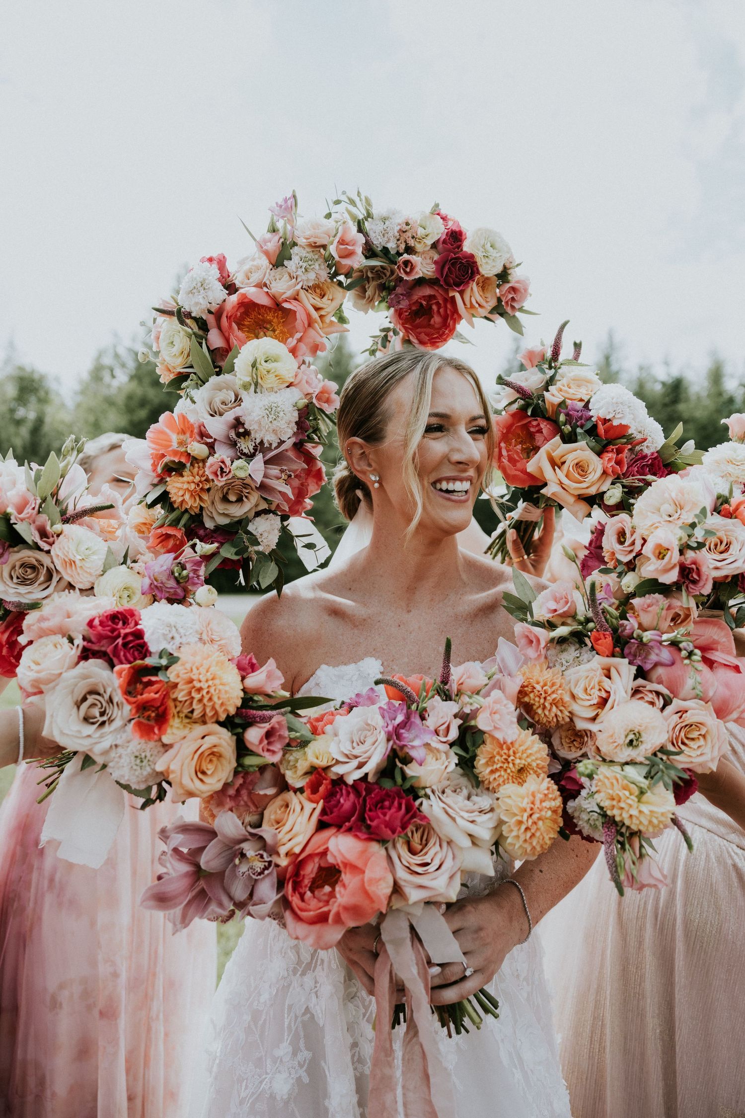 A stunning bridal bouquet featuring coral, peach and pink roses with lush greenery against a soft outdoor background.