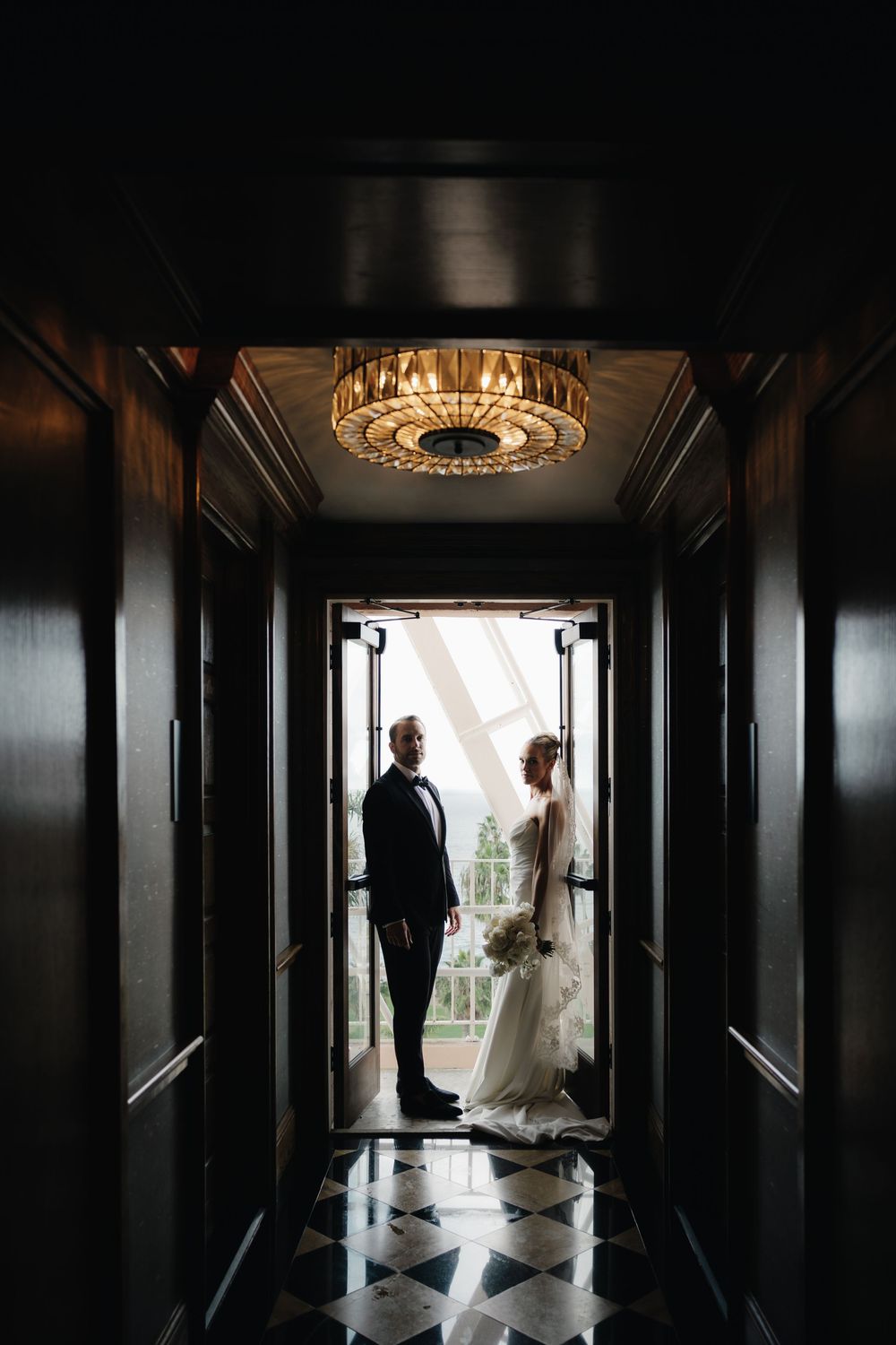 Bride and groom silhouetted in elegant dark hallway with chandelier and checkered marble floor.