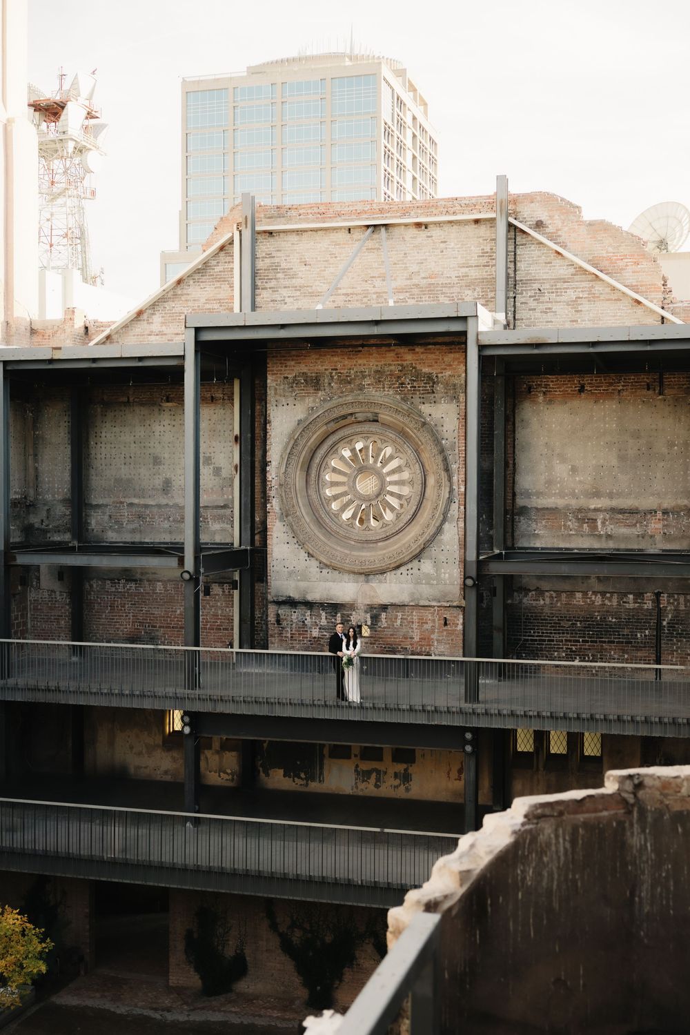 Wedding couple standing on a balcony of a historic brick building with ornate rose window in urban setting.