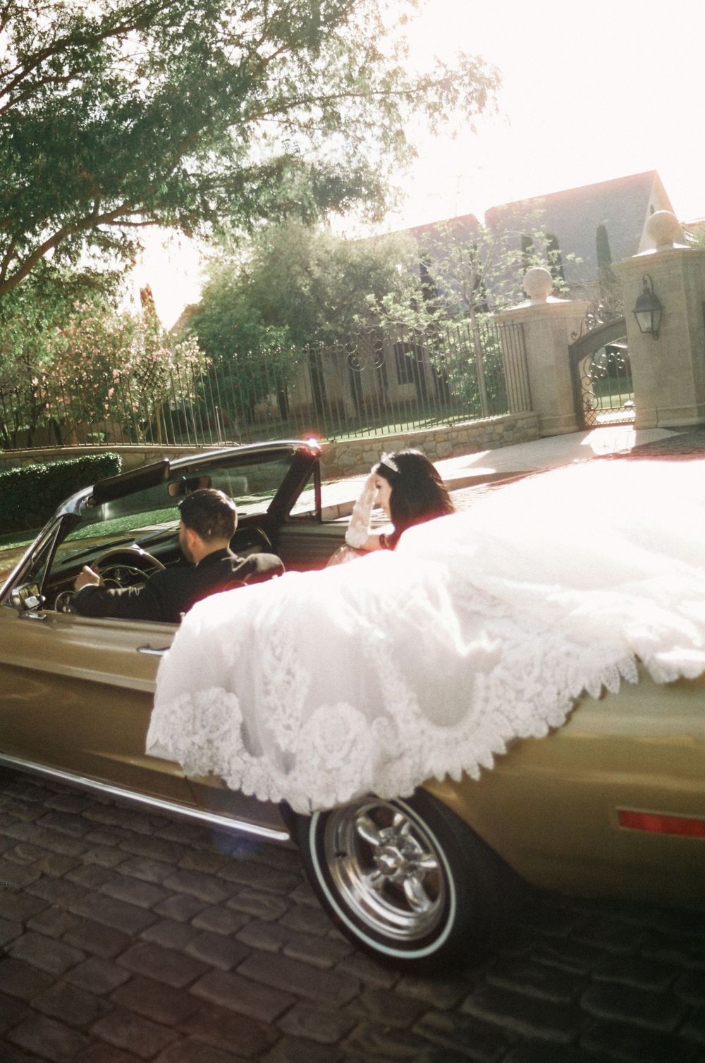 Bride in white lace gown sits with groom in a gold vintage convertible Mustang on cobblestone drive outside elegant estate.