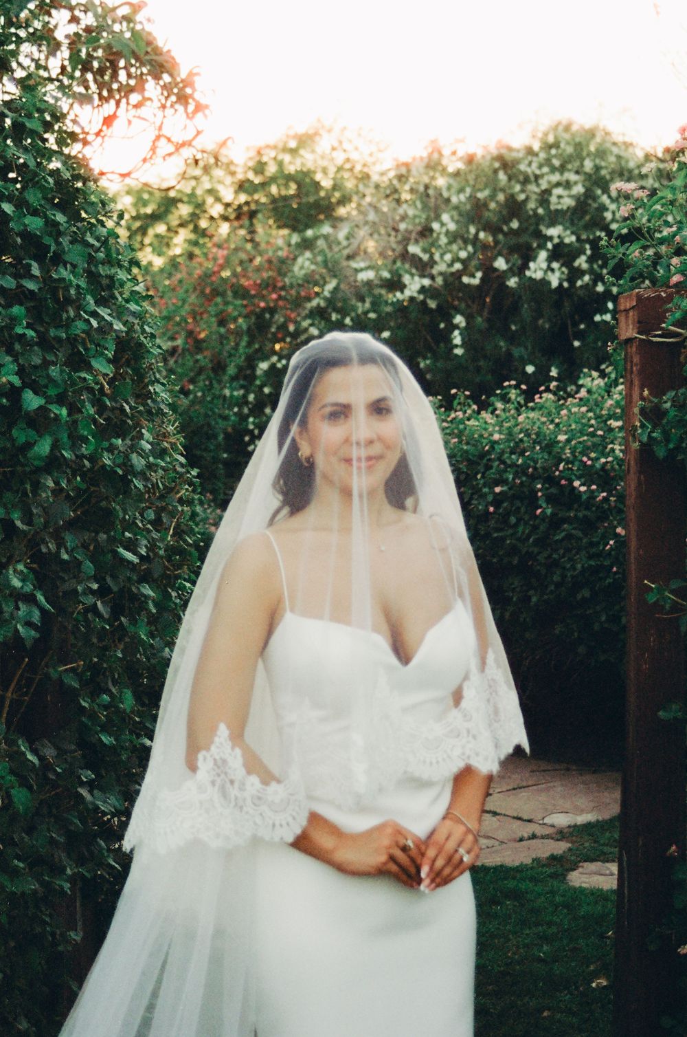 Bride in white gown and lace-trimmed veil stands in lush garden pathway at golden hour.