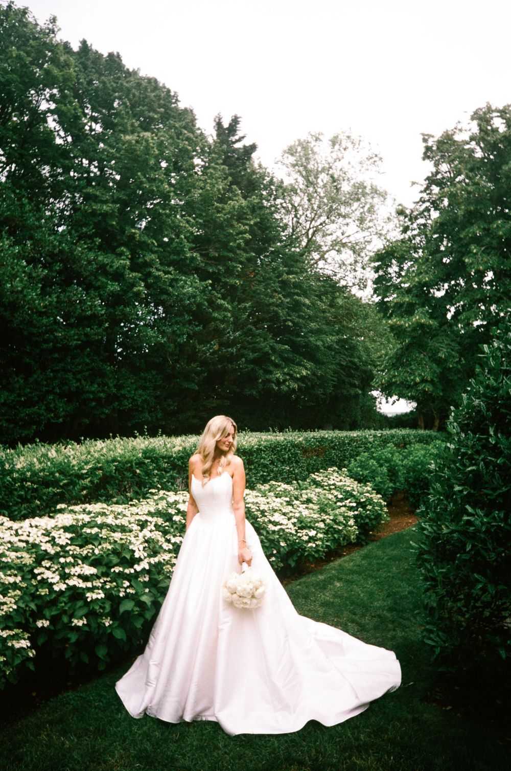 Bride in white ballgown holding bouquet stands in lush garden path surrounded by green hedges and white flowers.