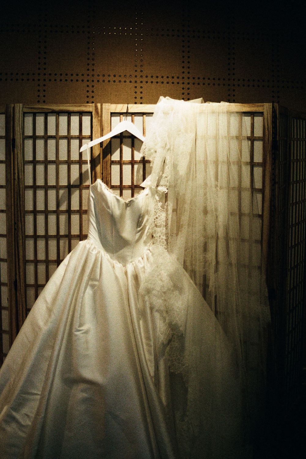 Elegant white wedding gown with lace details hanging on a wooden hanger against a decorative folding screen backdrop.