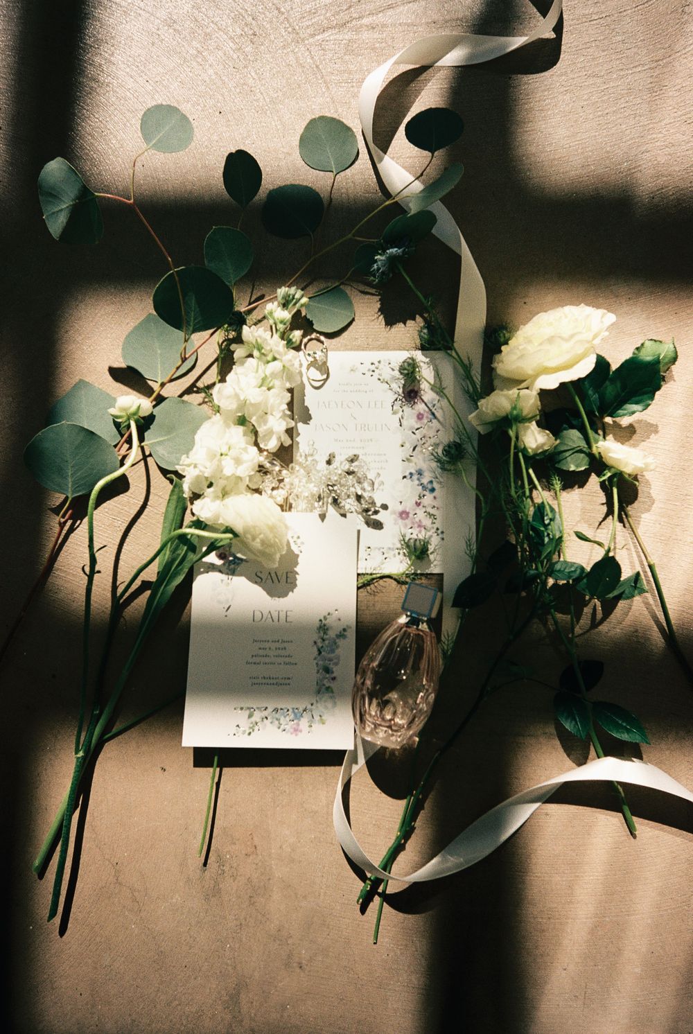 Elegant wedding flat lay with white flowers, eucalyptus, ribbon, invitation cards, and perfume on a neutral background.