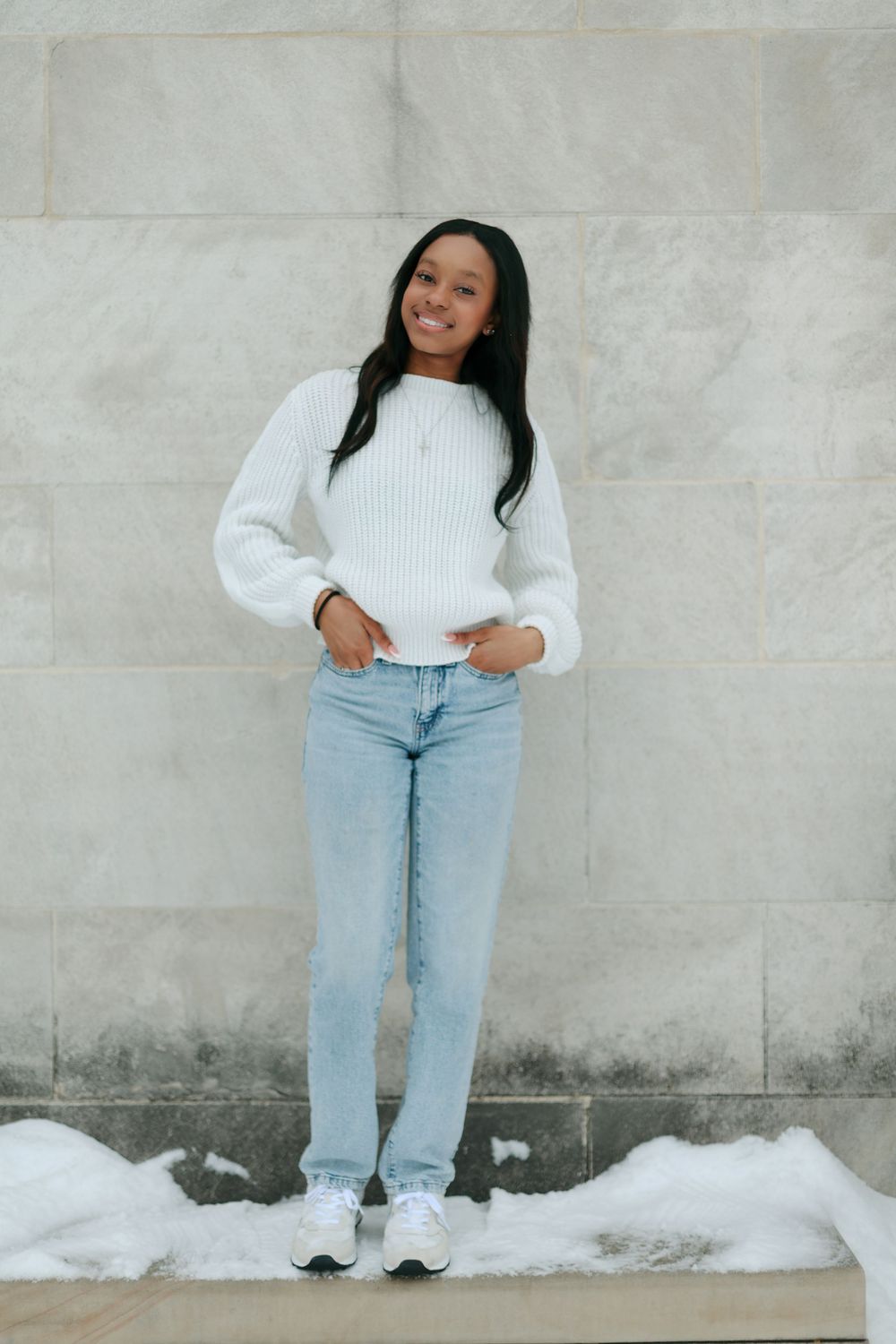 A person in casual winter attire poses against a concrete wall with snow on the ground.