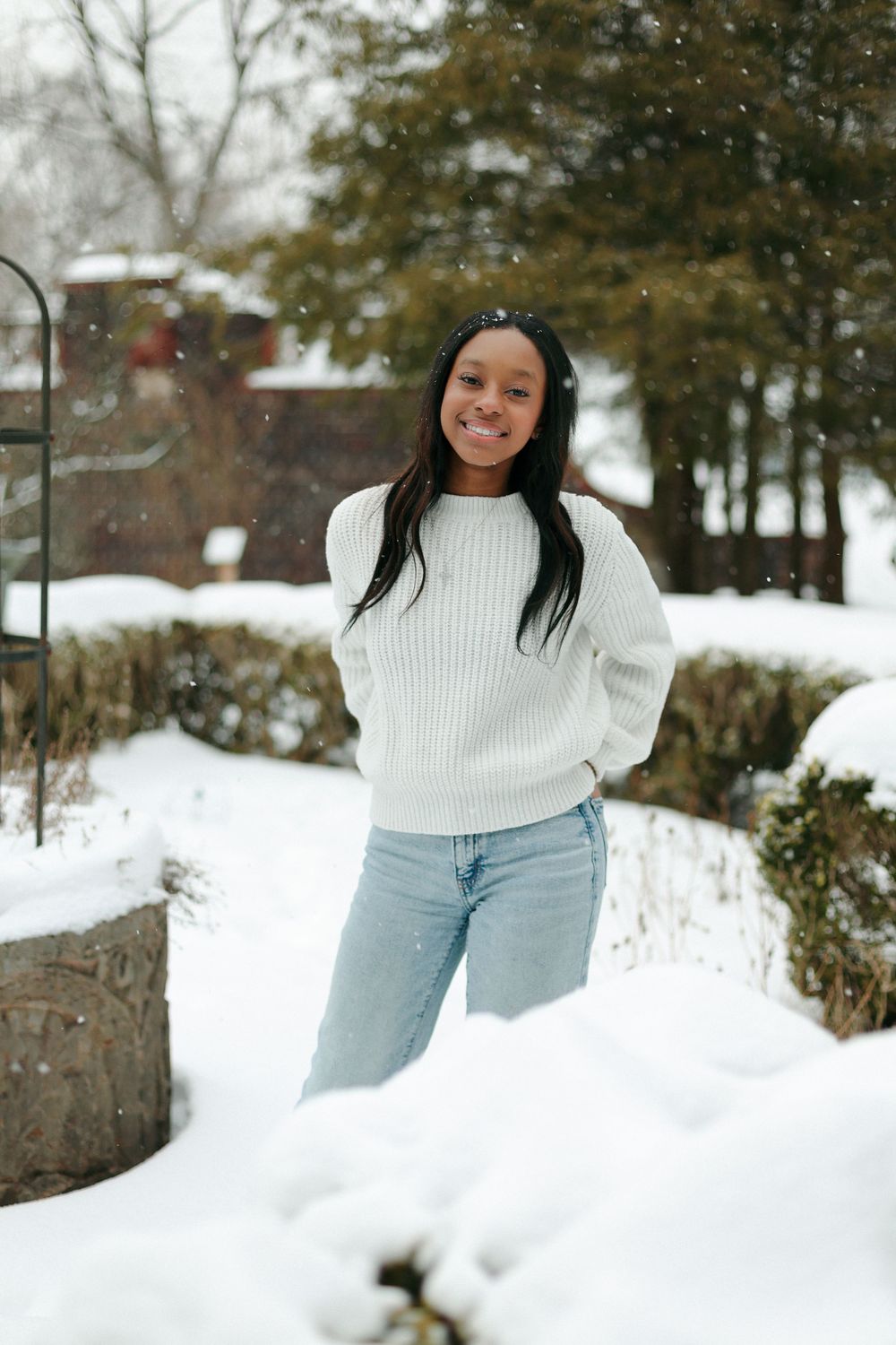 Someone in a cozy white sweater and light jeans poses cheerfully in a snowy winter landscape.