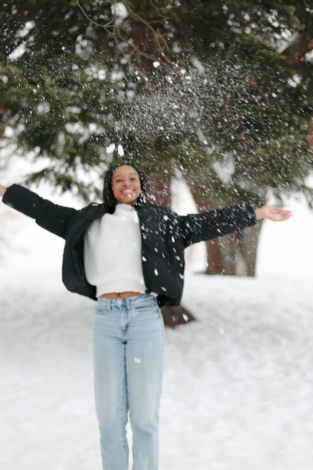 A person wearing light jeans and dark top plays joyfully in deep winter snow beneath evergreen trees.