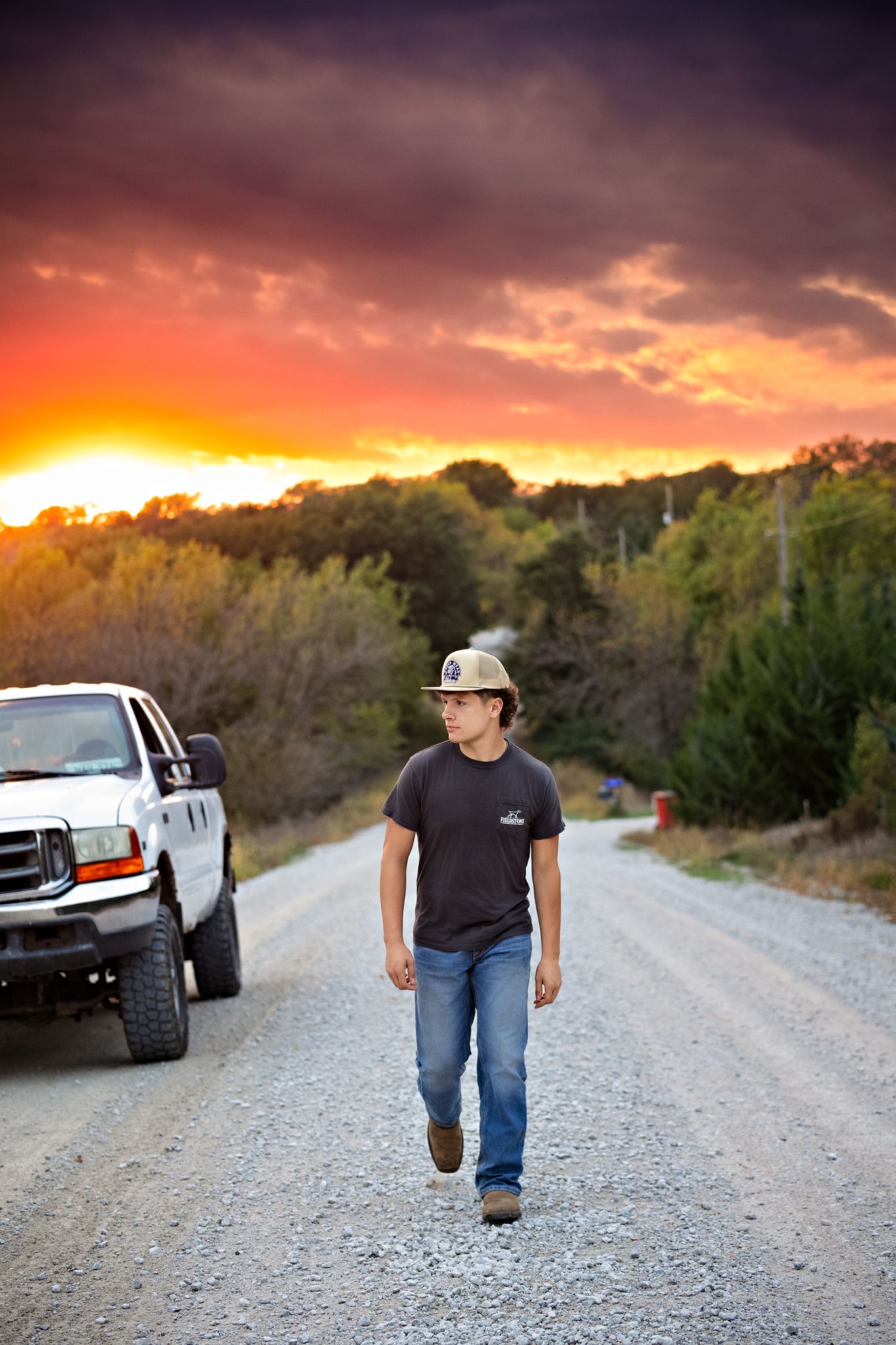 A person walks on a gravel road at sunset with dramatic orange clouds in the sky and trees silhouetted in the background.