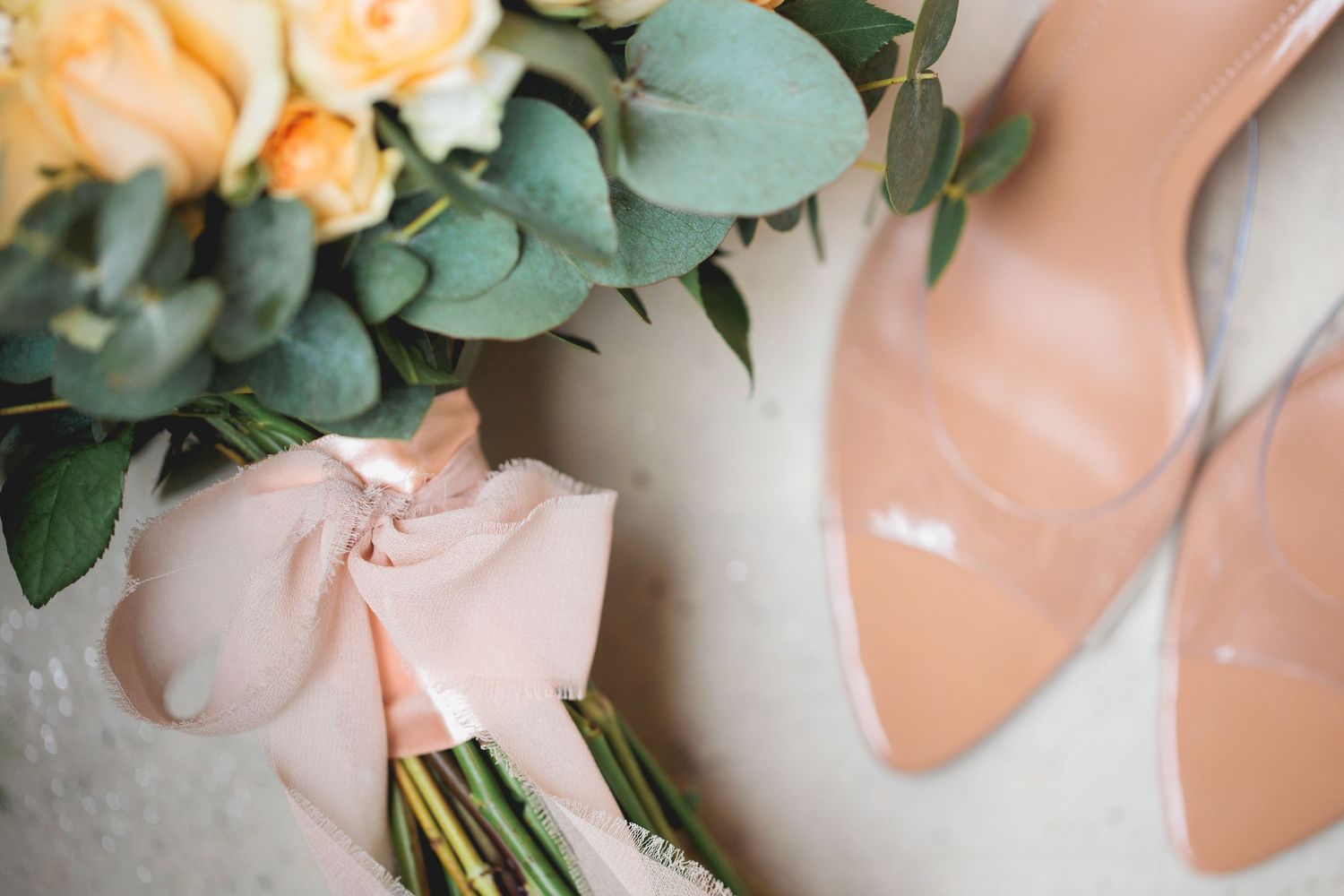 A bouquet of peach roses with eucalyptus leaves rests beside blush pink high heeled shoes.