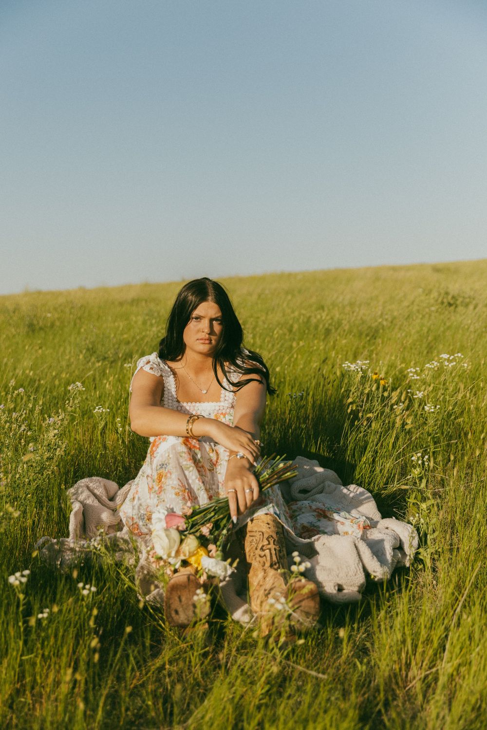 A person in a floral dress reclines in a sunlit meadow filled with tall grass and wildflowers on a summer day.